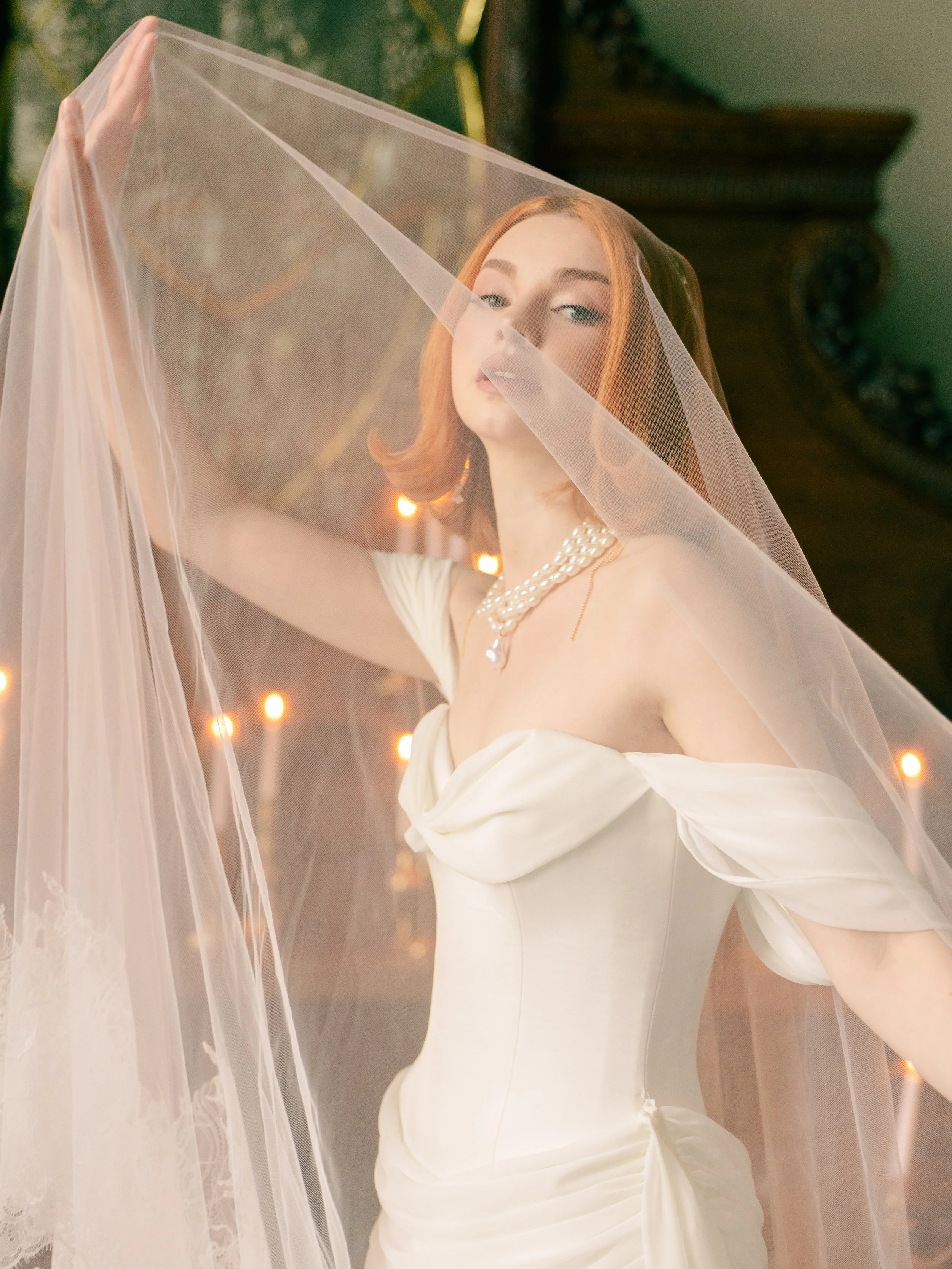 A bride with long red hair, wearing a white wedding dress and pearl necklace, holding a sheer veil over her face with warm lighting in the background.