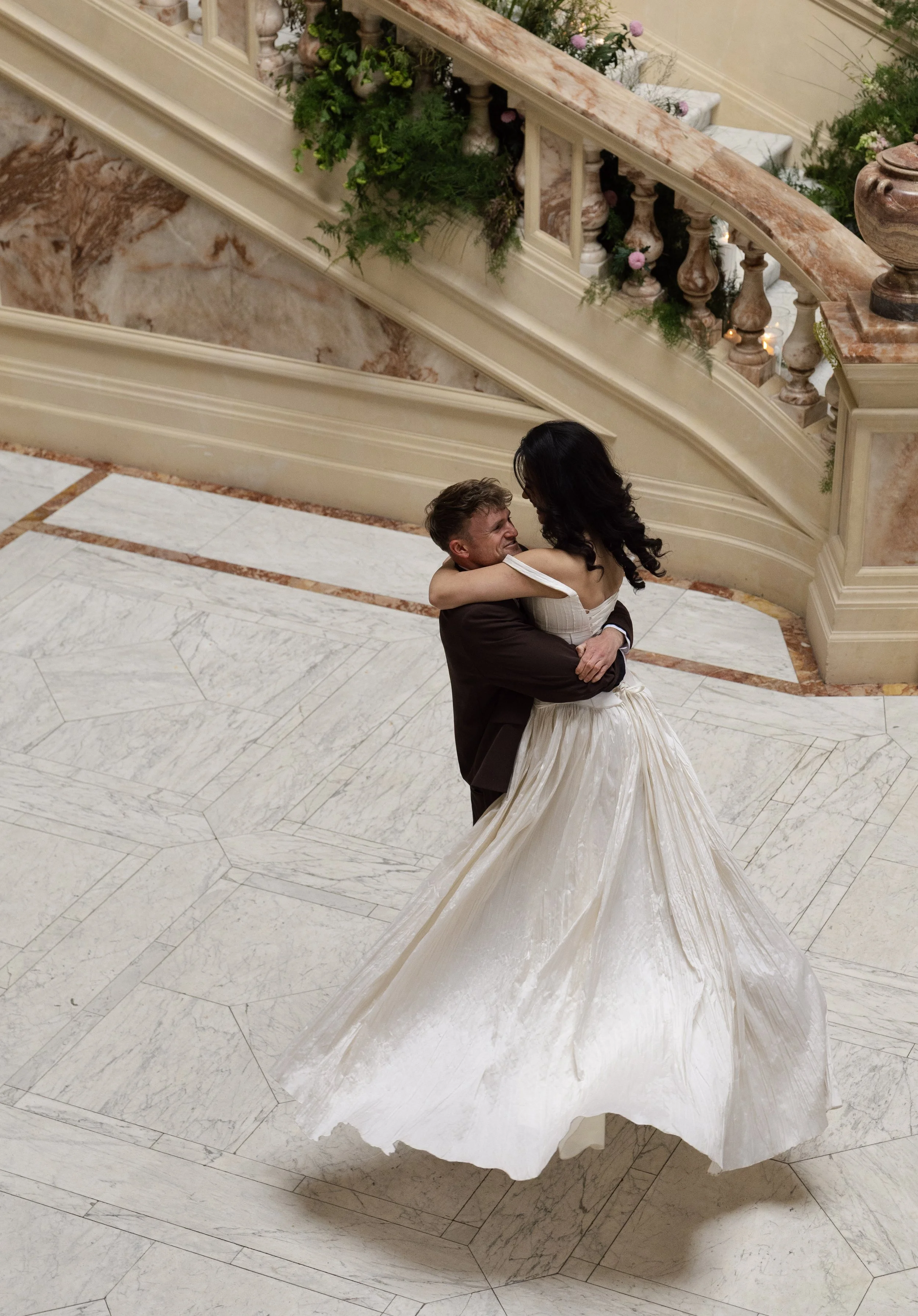 A man and woman are dancing closely on a marble floor in an elegant setting, with the woman in a white wedding gown and the man in a dark suit.