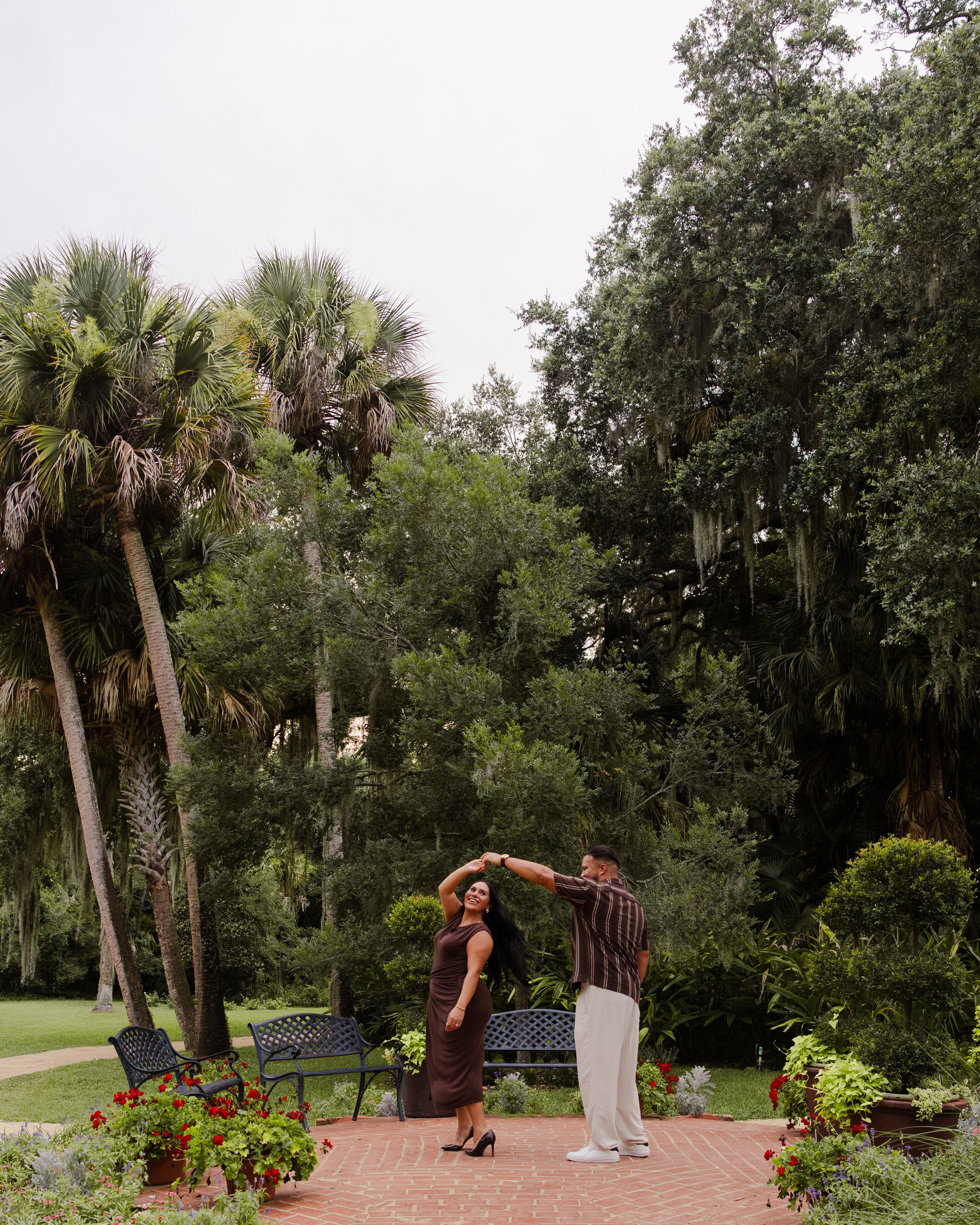 A man and woman dancing outdoors on a brick patio, surrounded by lush green trees and colorful flowers, with black garden benches nearby.