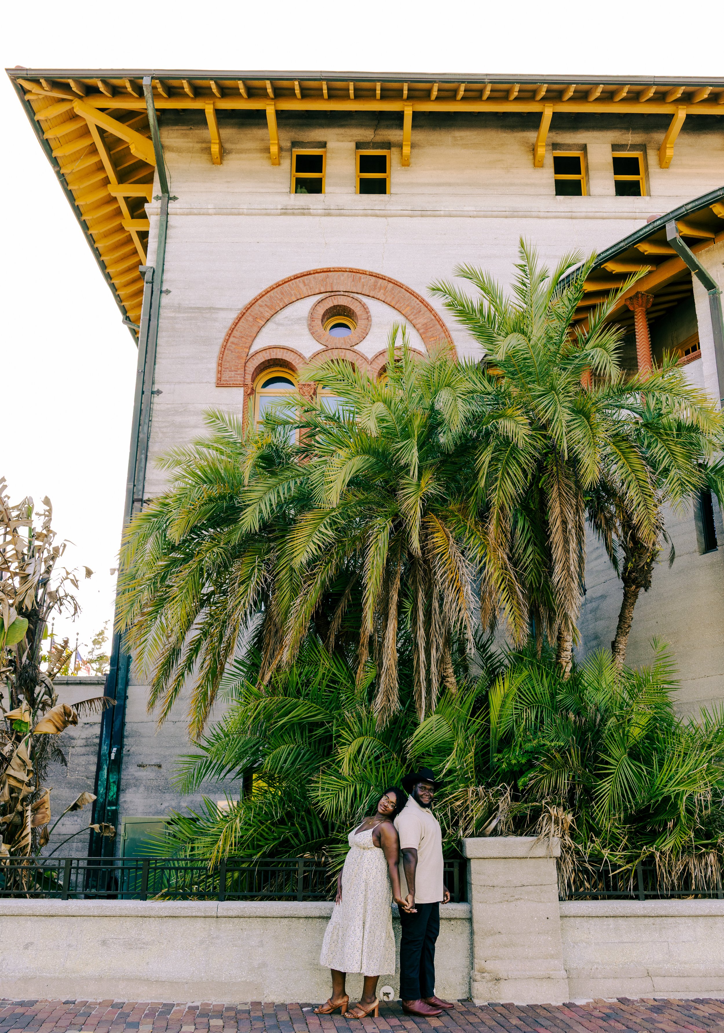 A couple stands hand in hand in front of a large building with lush palm trees, brick accents, and yellow window frames during daytime.