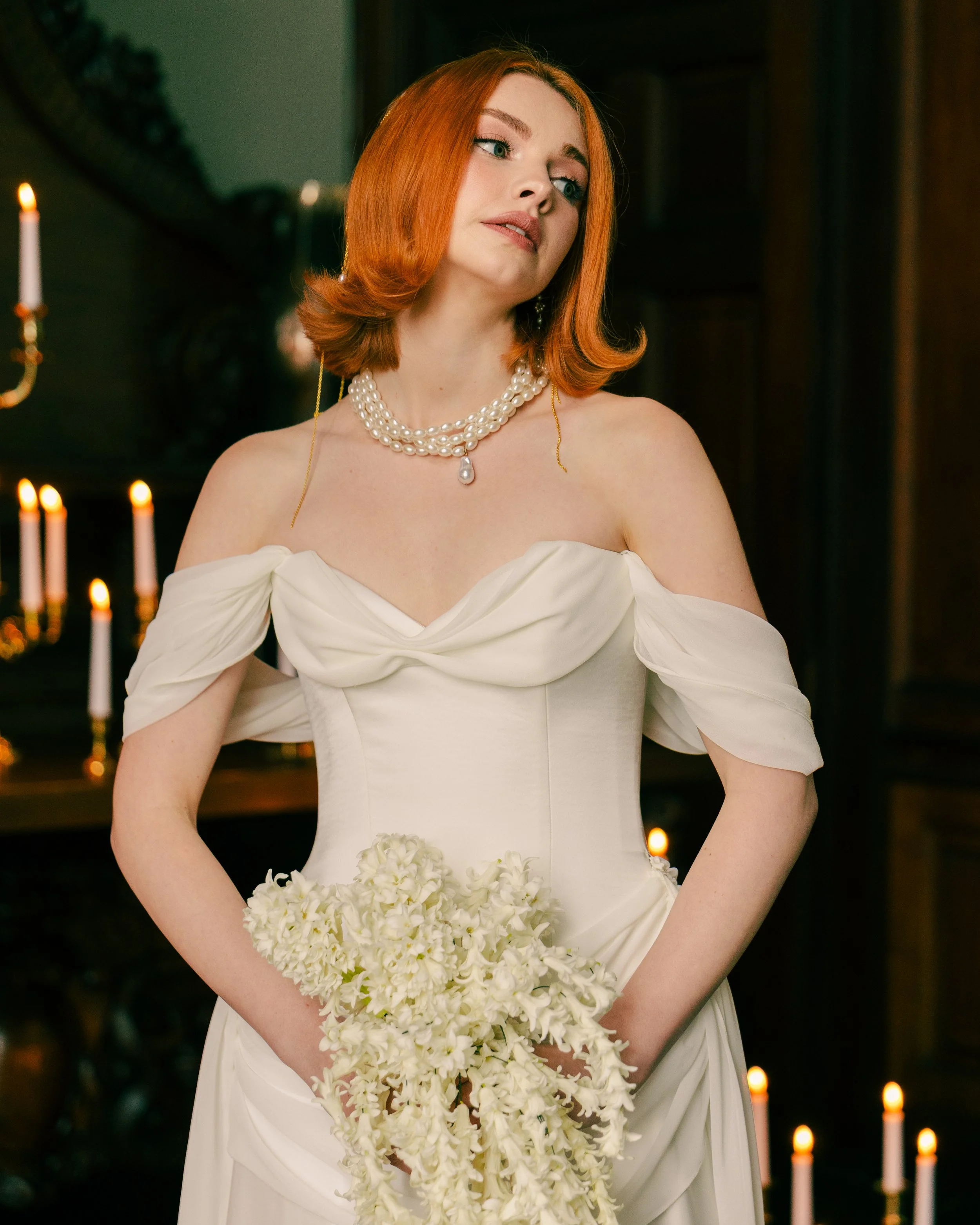 A woman with red hair in a white wedding dress holding a bouquet of white flowers, standing in front of lit candles.