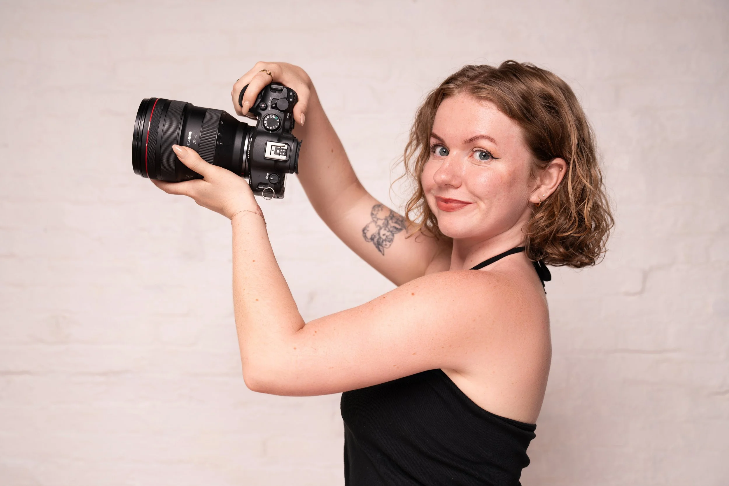 A young woman with curly red hair and a tattoo on her arm holds a professional camera, slightly smiling, against a plain white wall.
