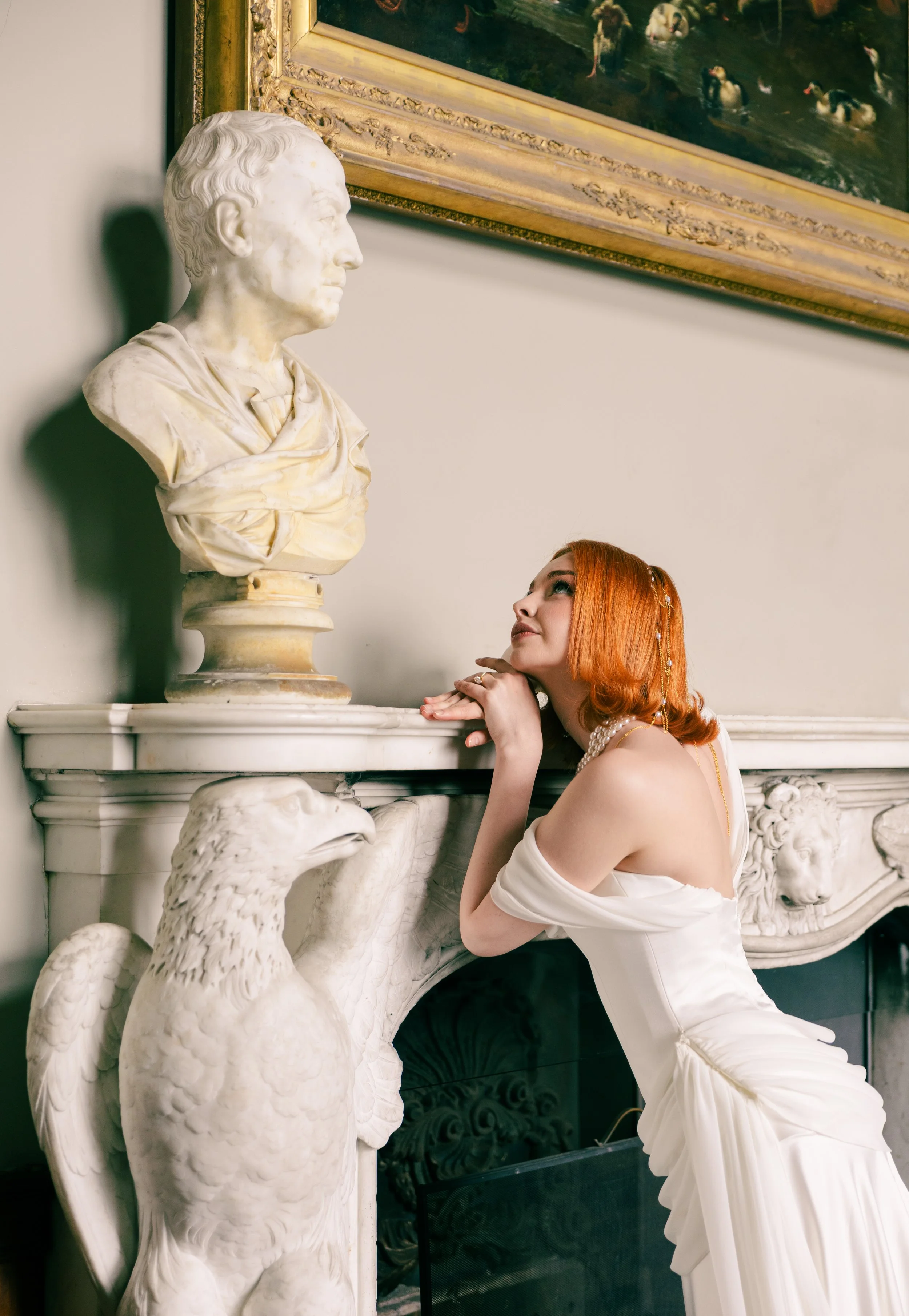 A woman with red hair, dressed in a white gown, sits beside a marble sculpture of a bust and a marble fireplace with eagle carvings, in an elegant room with a painting of ducks on the wall.
