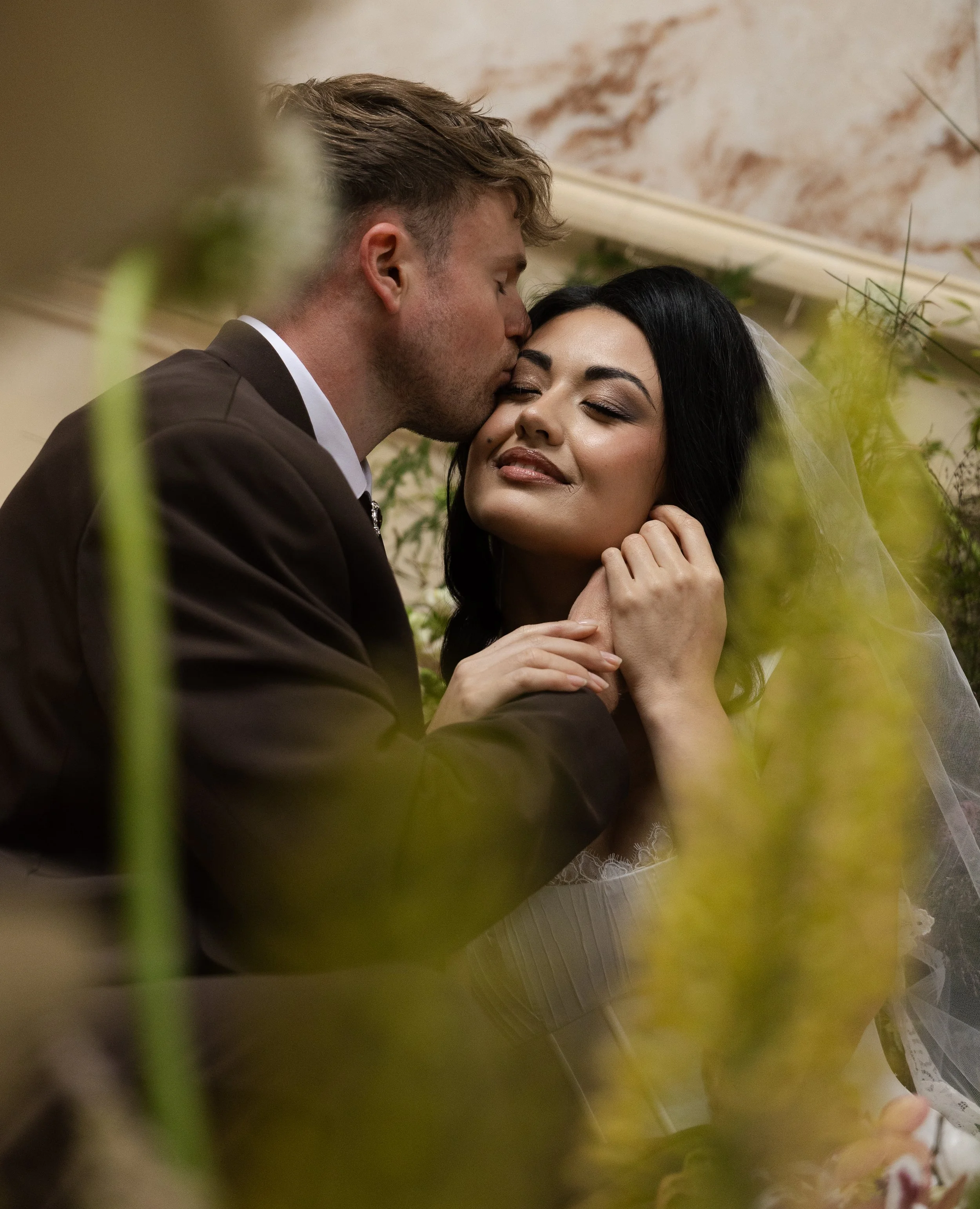 A groom kisses a bride on the forehead during their wedding ceremony, with greenery in the foreground and a marble wall in the background.