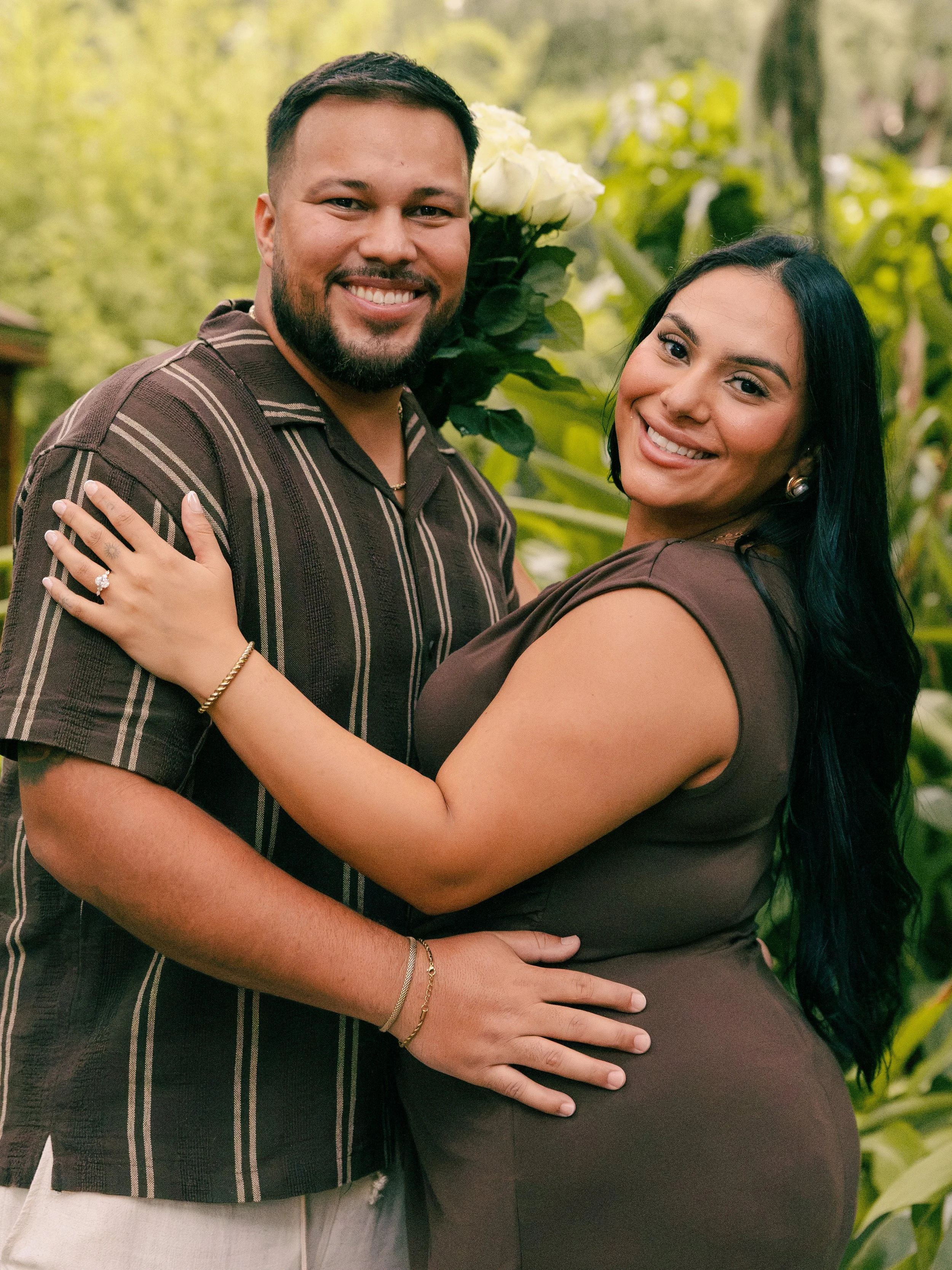 A couple standing outdoors, smiling, with the woman holding a bouquet of white roses behind their heads. The woman is pregnant, wearing a fitted dress, and the man is wearing a striped shirt. They are embracing, with the woman's hand resting on the m