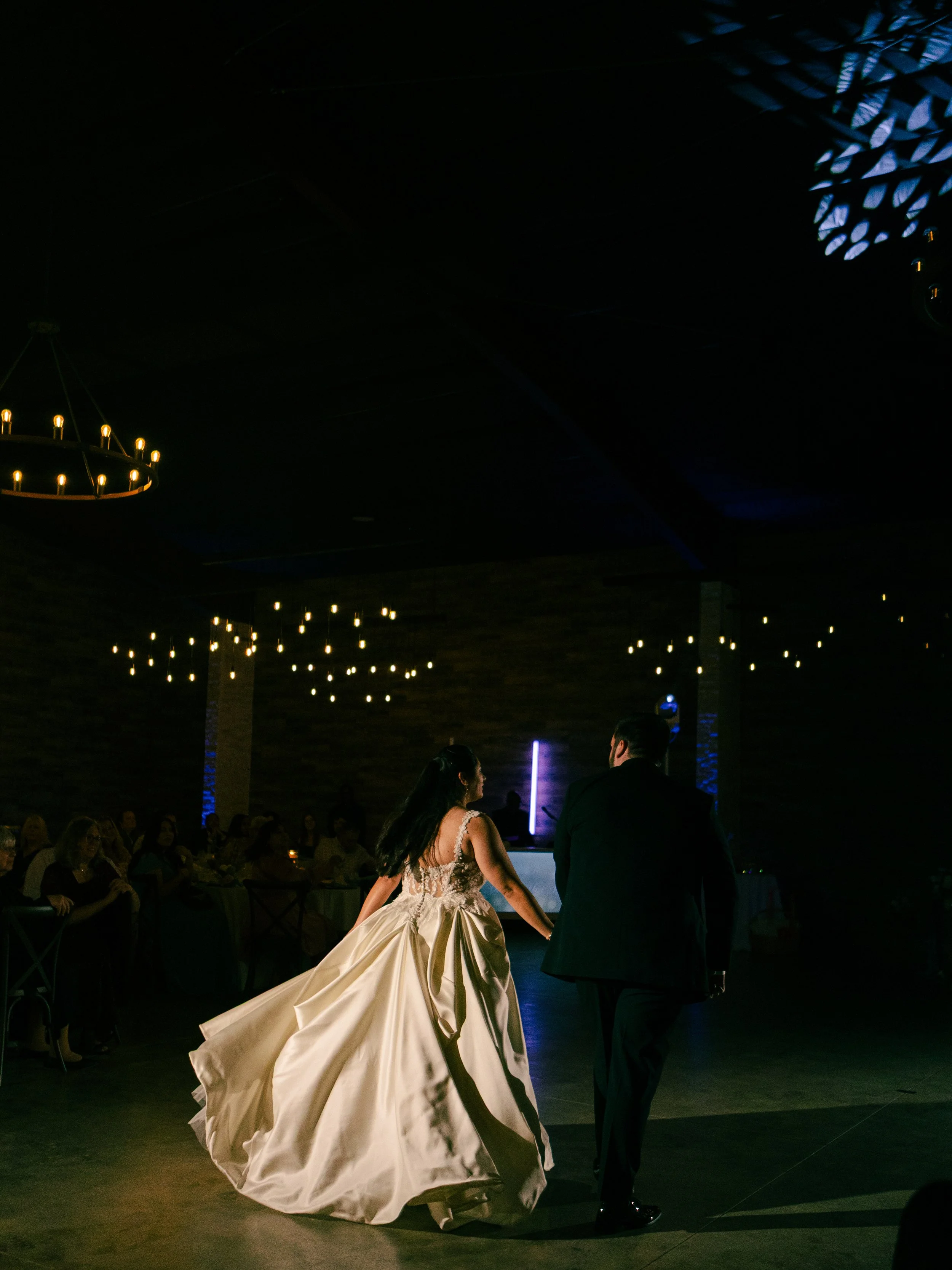 A bride in a wedding dress and a groom in a black tuxedo dancing at their wedding reception with guests seated at tables in the background, illuminated by hanging string lights and chandeliers.