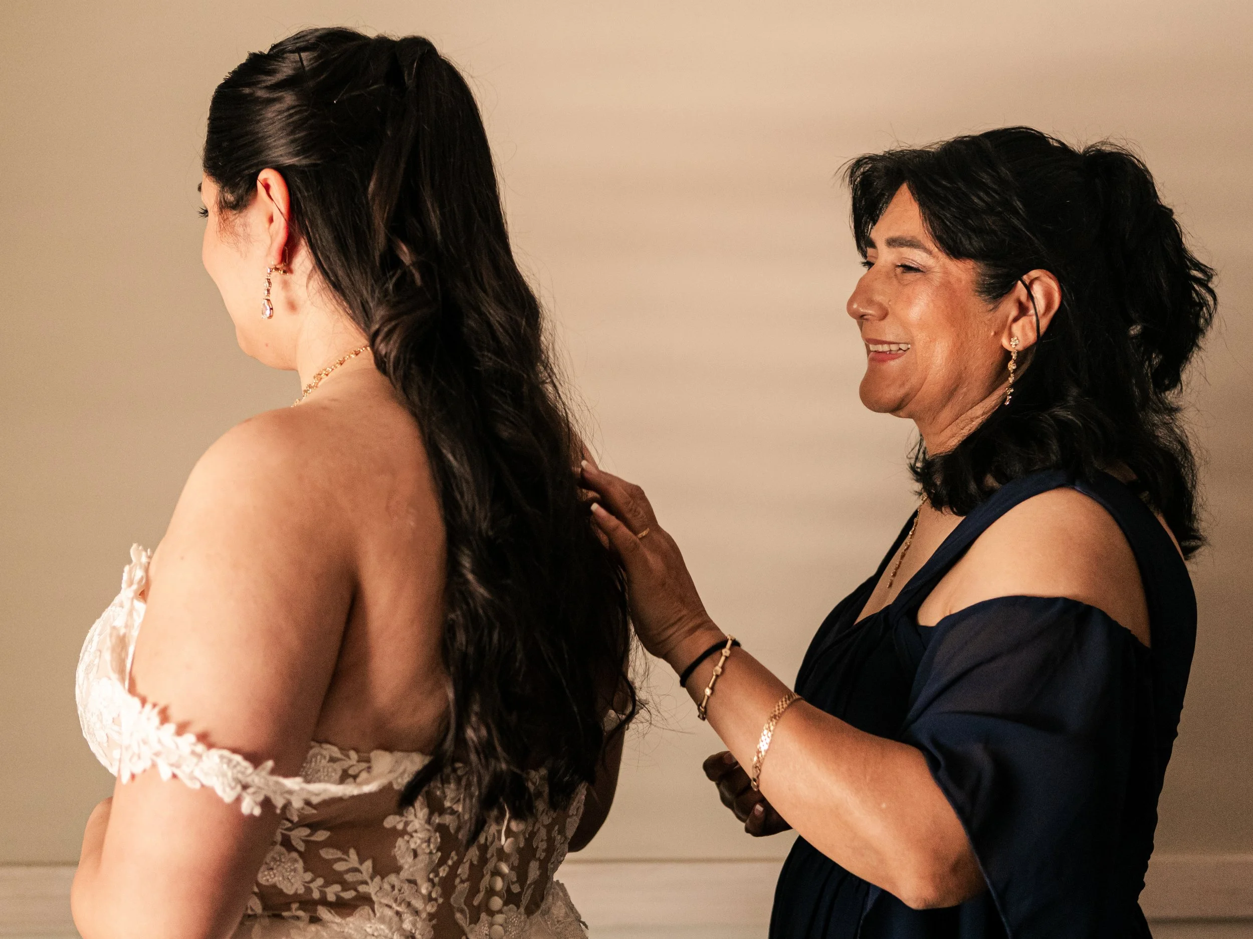 A woman adjusting the back of another woman's wedding dress as they smile and interact with each other.