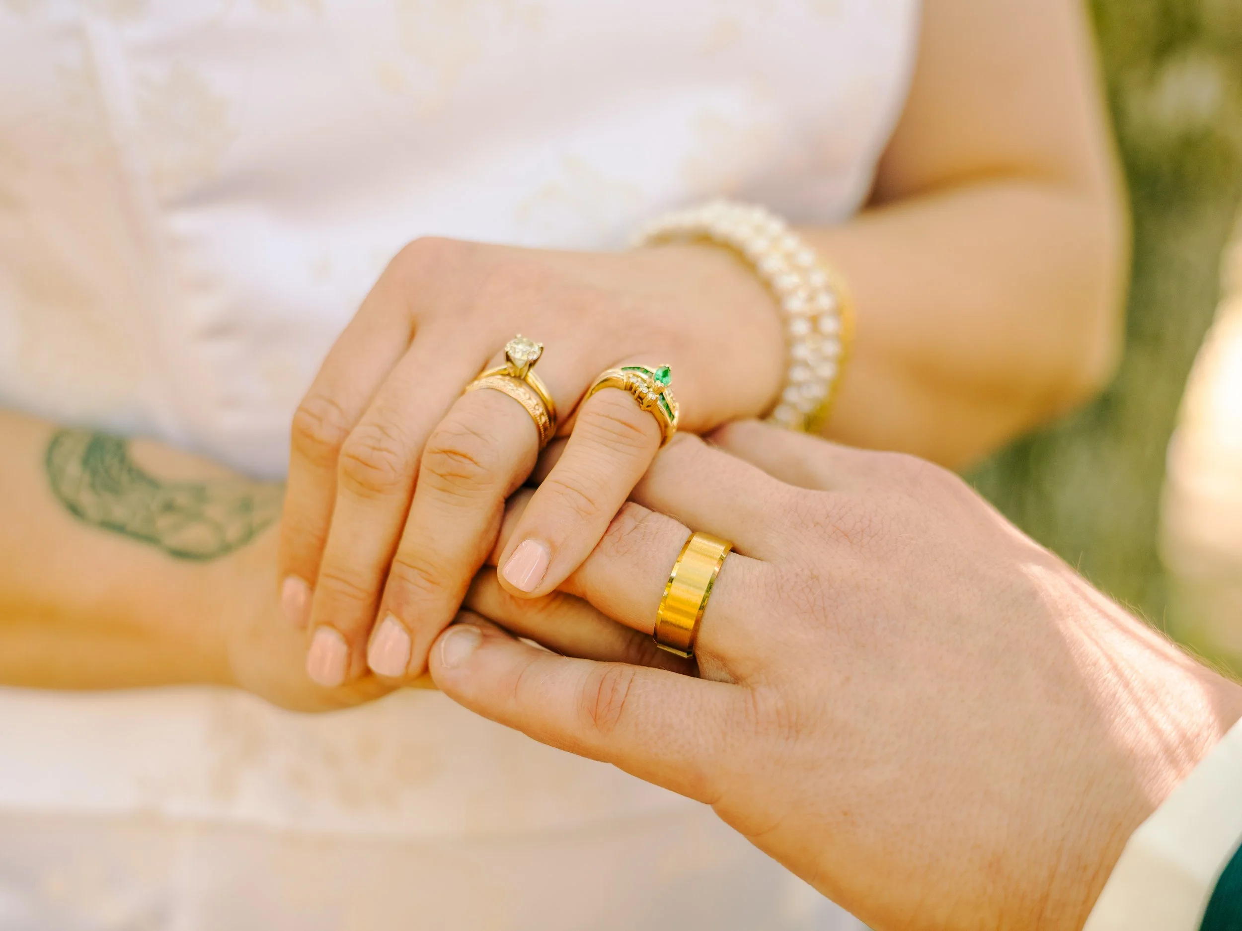 Close-up of two hands embracing, showing multiple gold rings with gemstones, one featuring a diamond and the other with an emerald, and a pearl bracelet on the wrist, with a tattoo visible on the arm and a white garment in the background.
