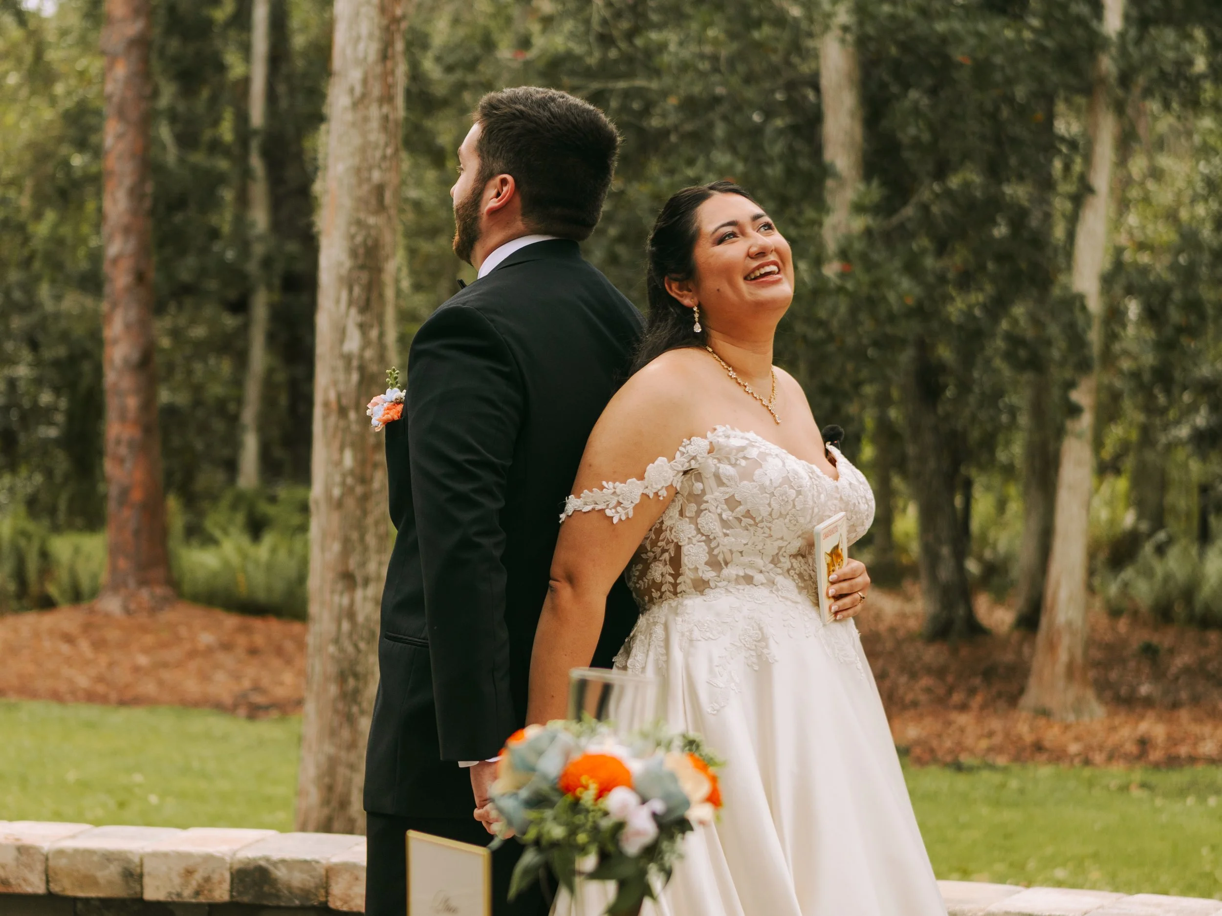 A bride and groom stand back-to-back outdoors during a wedding ceremony. The bride is smiling and looking upwards, wearing a white lace wedding dress with an off-shoulder design and jewelry. The groom, dressed in a black tuxedo with a boutonniere, is