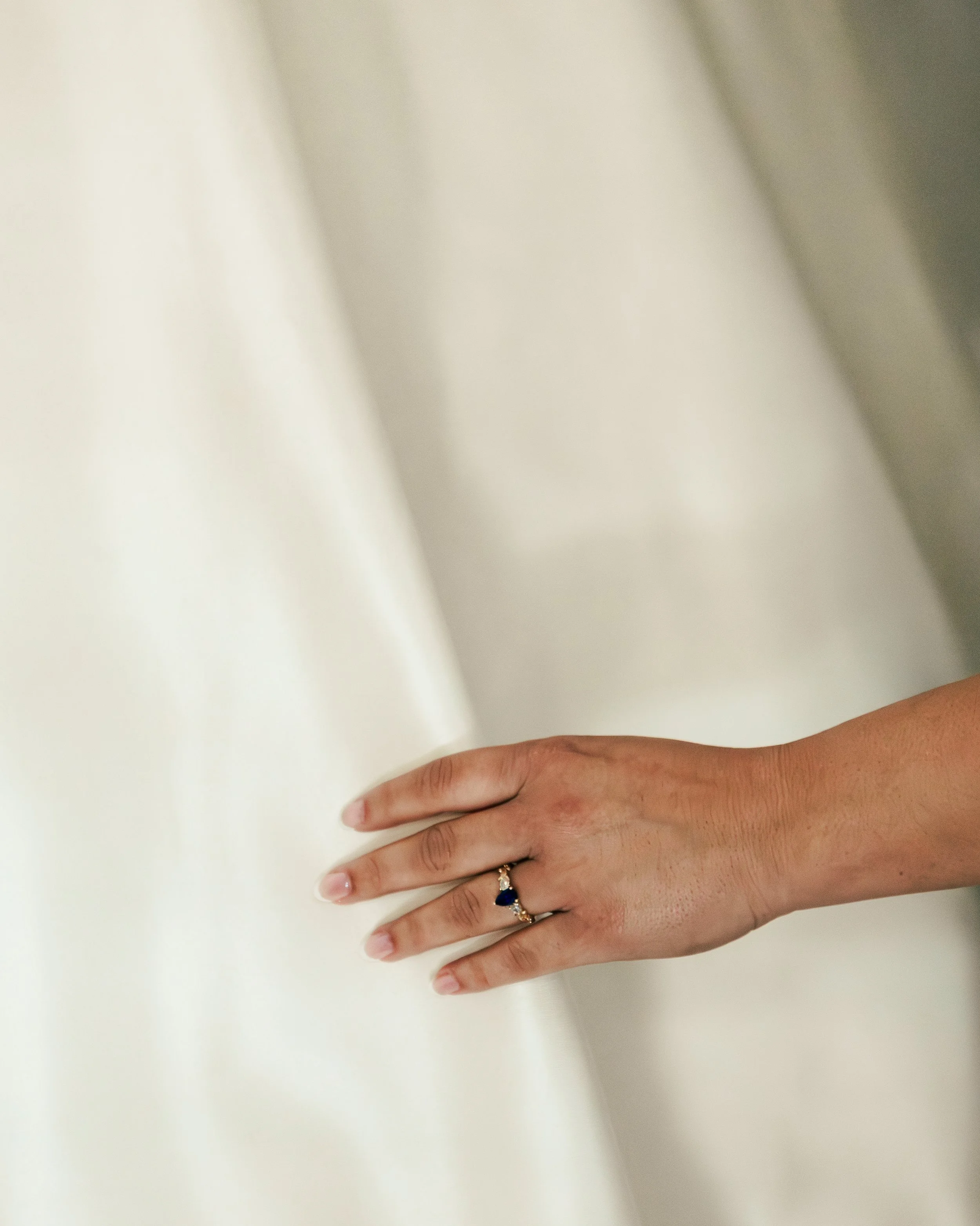 A close-up of a woman's hand resting on a white surface, wearing a ring with a dark blue gemstone and a wedding band.