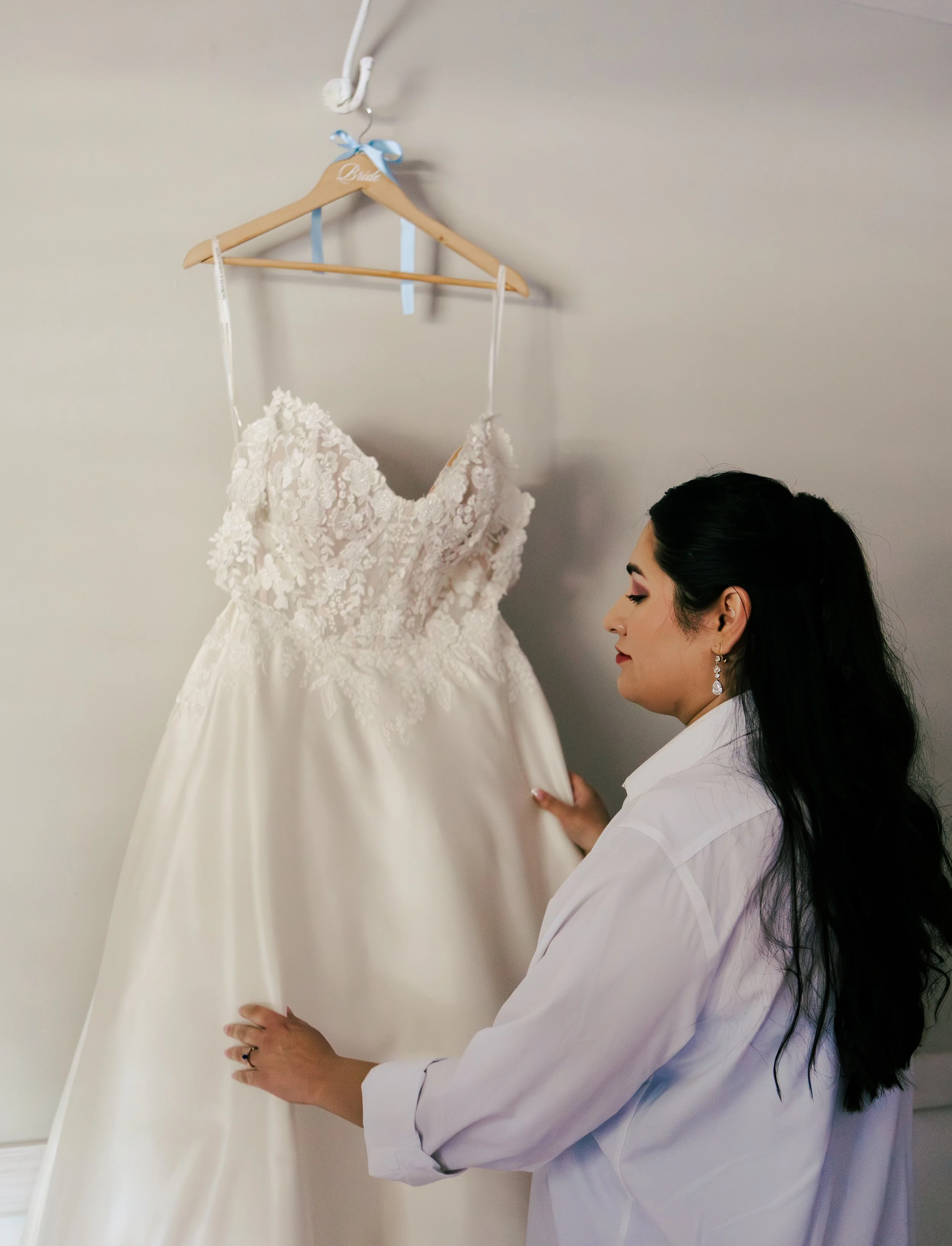 A woman with dark hair styled in waves and wearing earrings is adjusting a wedding dress hanging on a wooden hanger with a blue ribbon. The dress has lace details and floral embellishments.