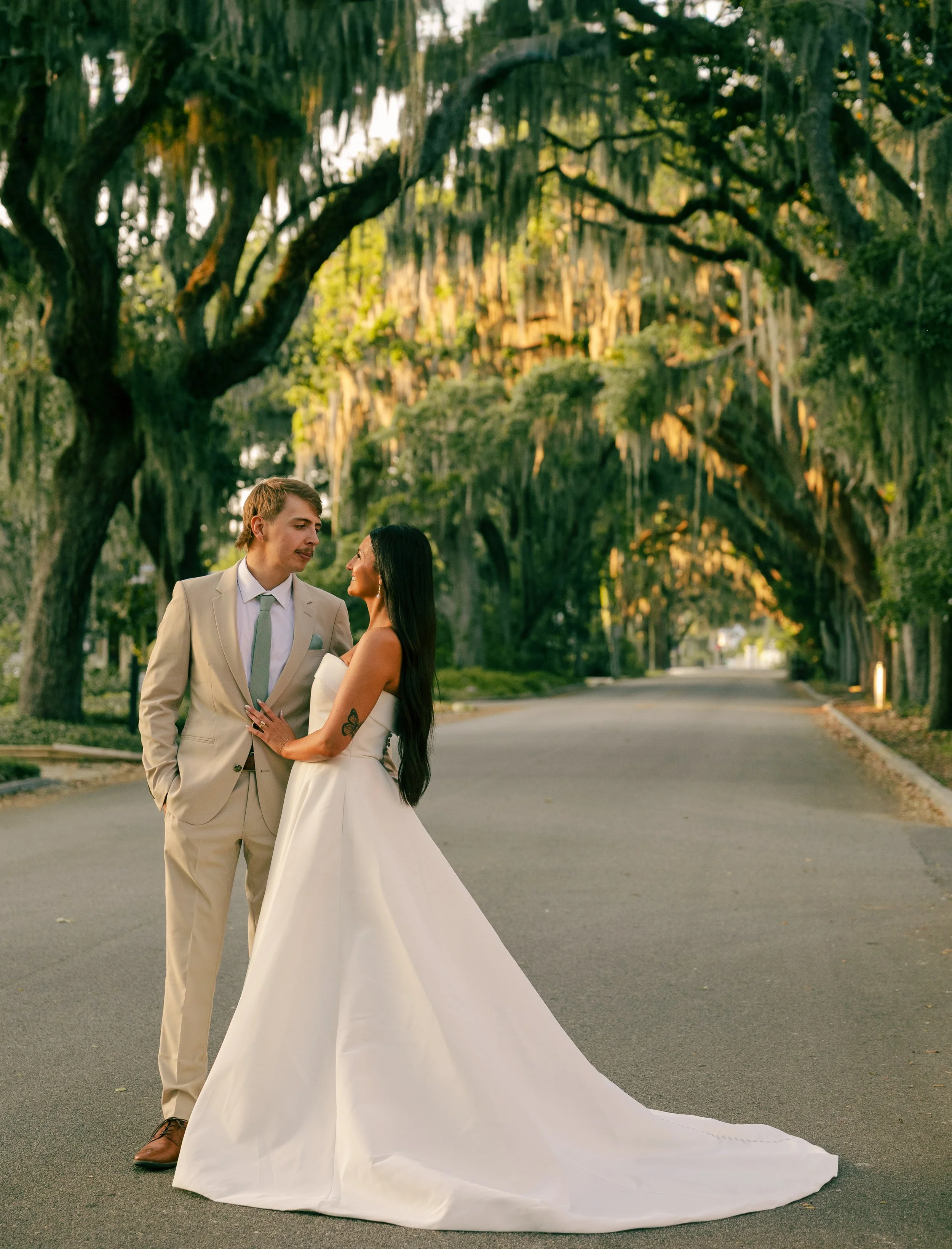 A couple dressed in wedding attire standing in an outdoor area with large, moss-covered trees, during what appears to be late afternoon or early evening.