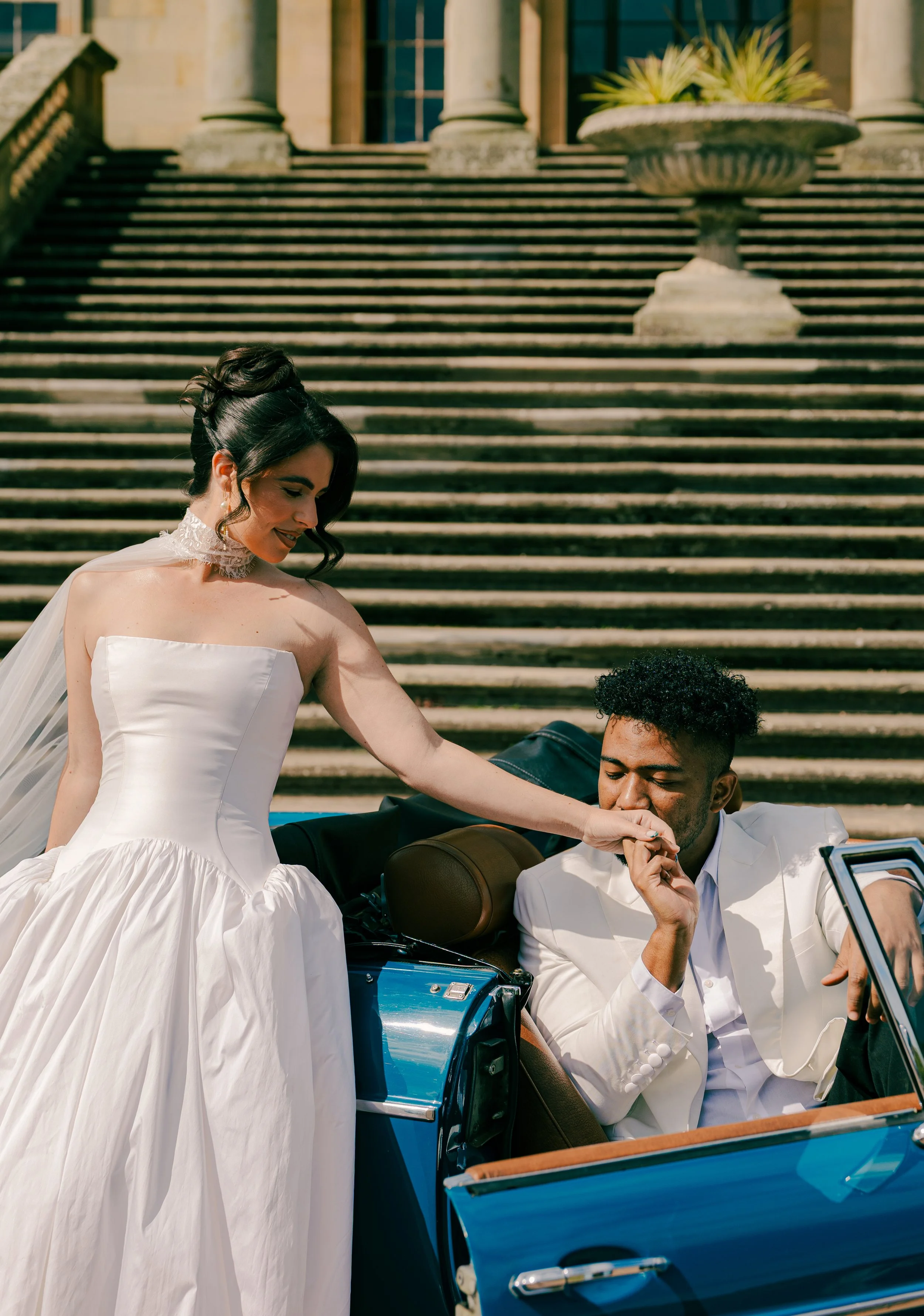 A woman in a wedding dress is leaning towards a man in a white suit who is seated in a blue convertible car, with the mall stairs and a stone planter visible in the background.