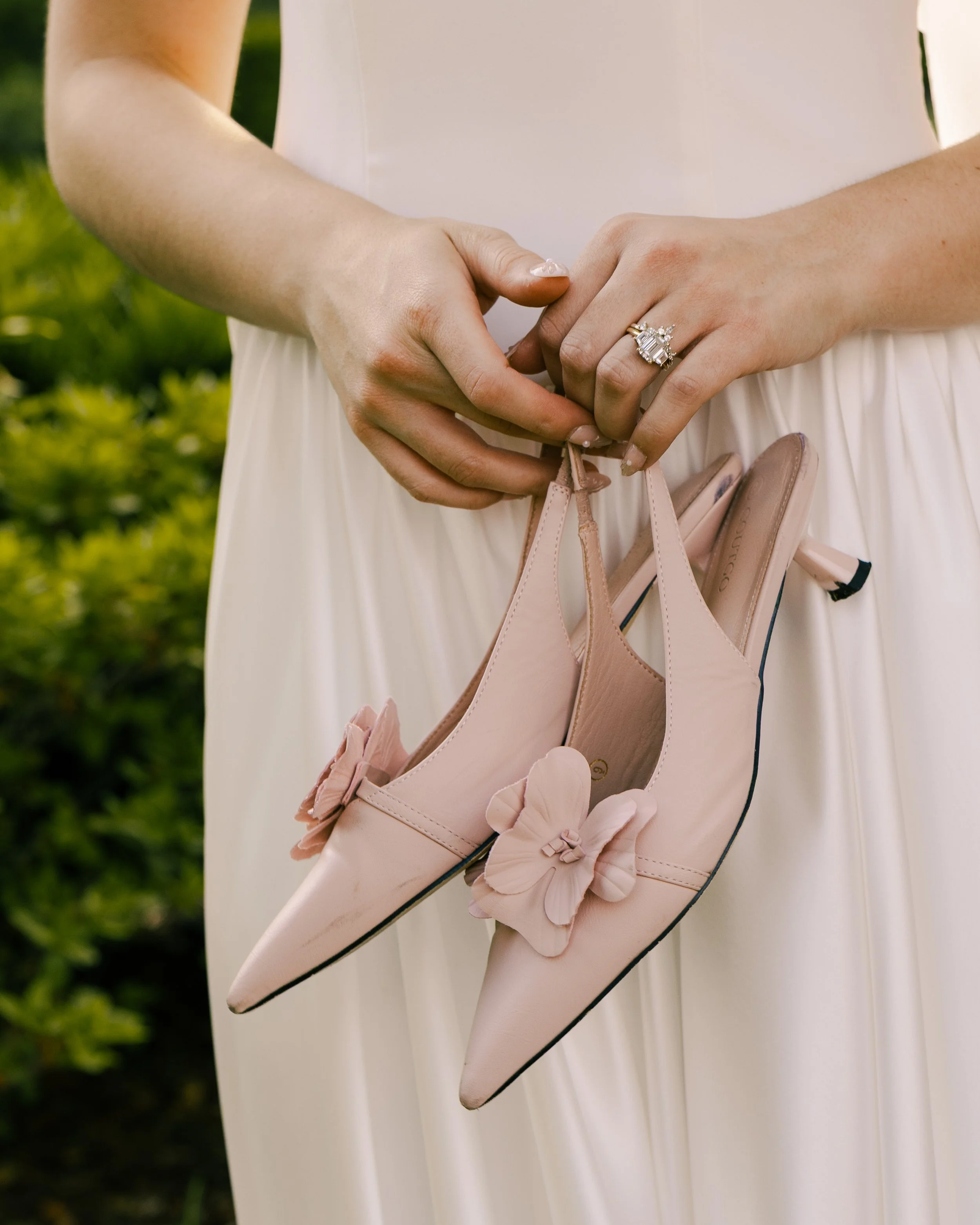 A bride in Jacksonville, Florida, holds pink kitten heels with a floral bow decal. She has a large square-cut engagement ring.