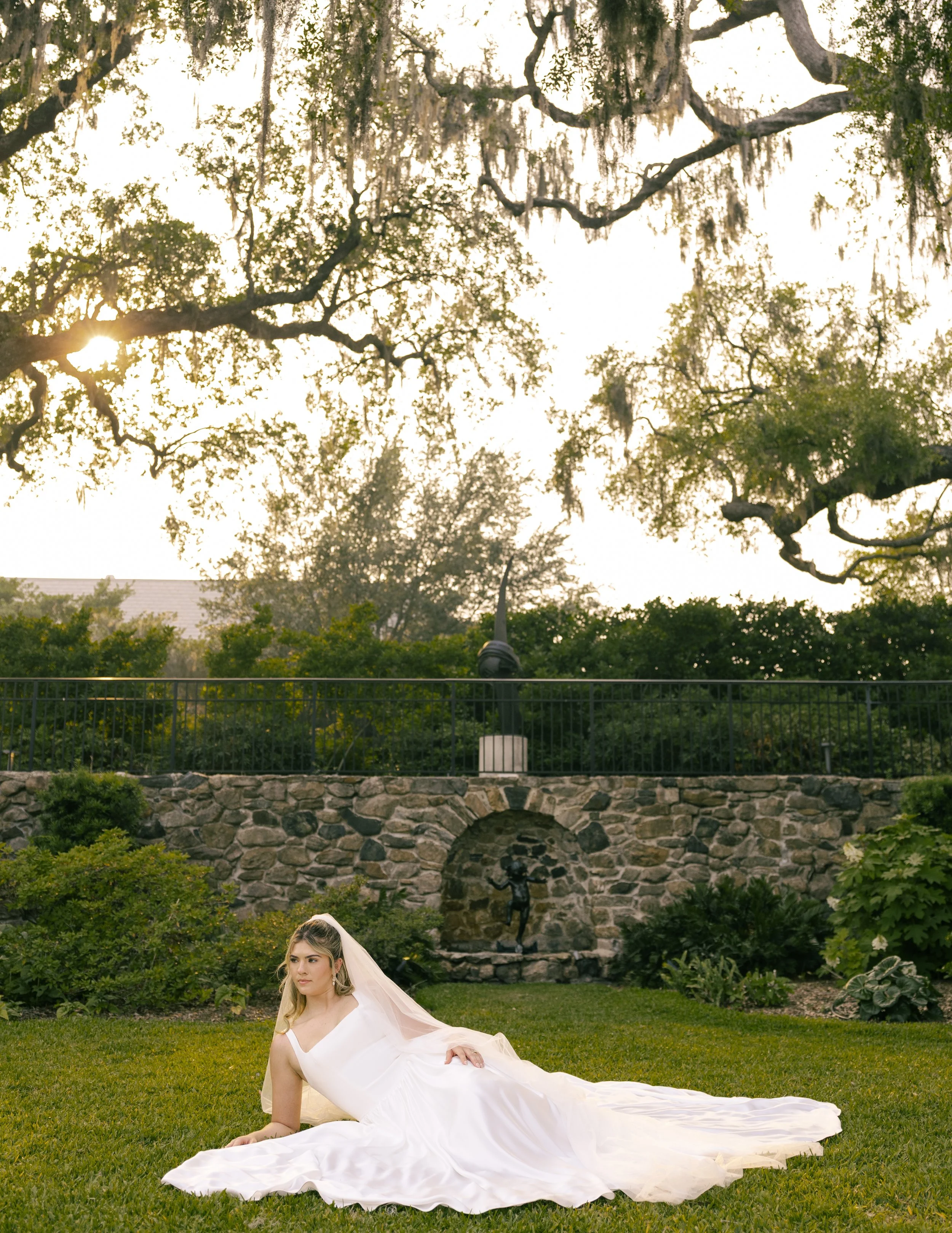 A bride in a white wedding dress lying on the grass in a garden, with a stone wall and trees in the background during daytime.