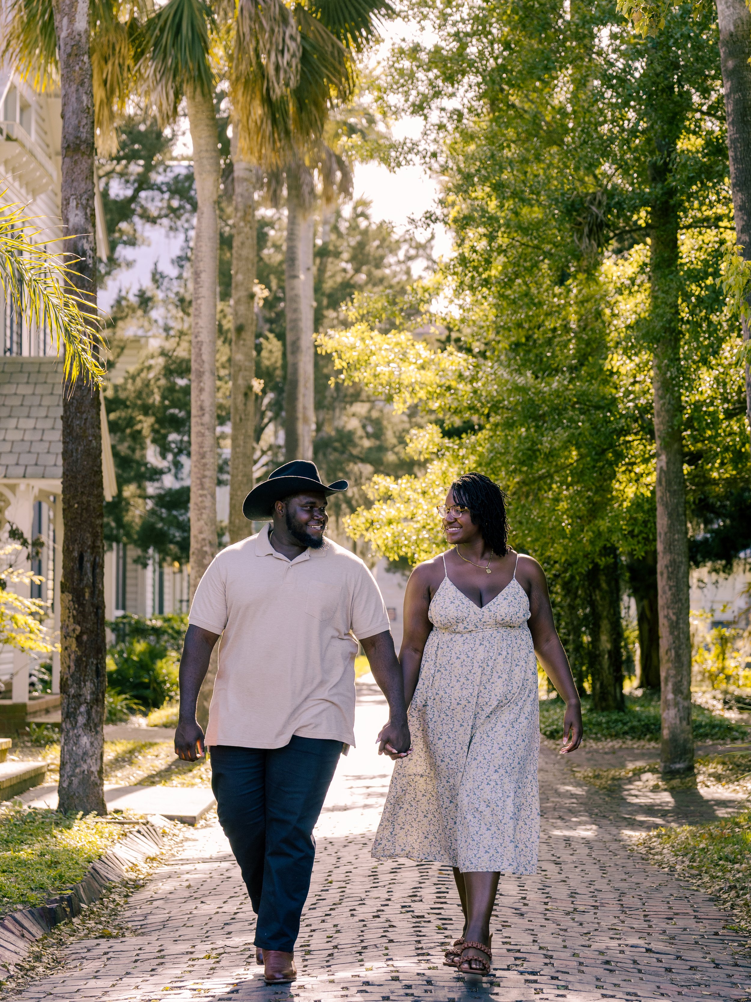 A man and woman walking hand in hand on a brick sidewalk through a park with green trees and sunlight filtering through the leaves, enjoying a warm day.