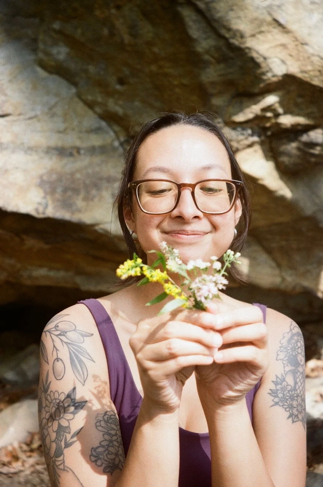 A woman with glasses and tattoos on her arms holding a small bouquet of flowers, smiling and looking down at the flowers, with a rocky background. Creative retreat host.