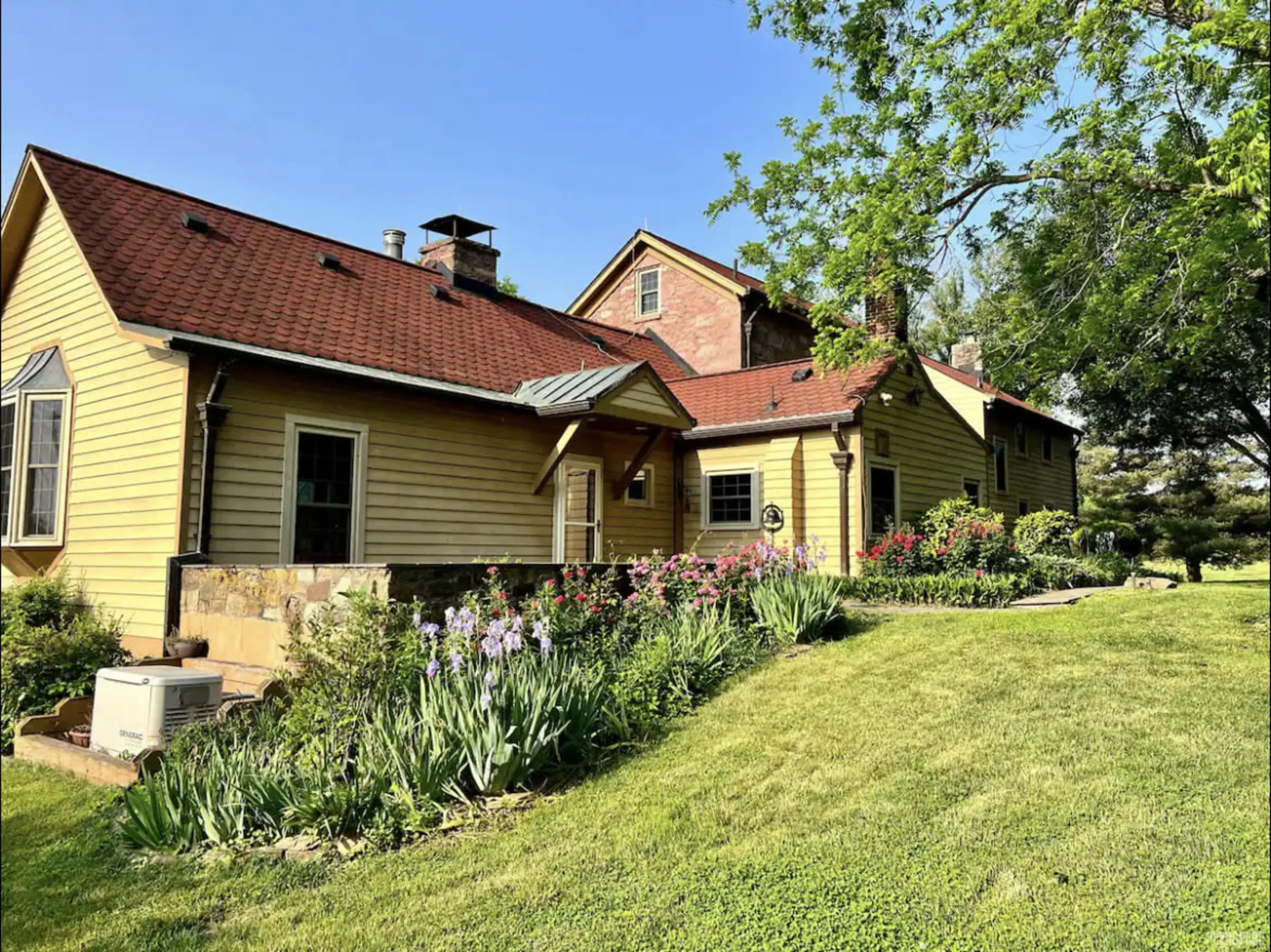 Farmhouse creative retreat location. Yellow house with red roof, surrounded by a well-manicured lawn and flower garden, under a bright blue sky with green trees.