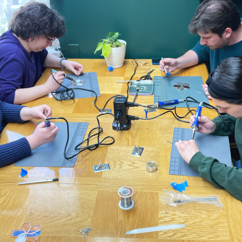 Craft workshop near Washington, D.C. Four people working at a wooden table on a soldering project, with tools, wires, and small metal parts. A potted plant is on the table.