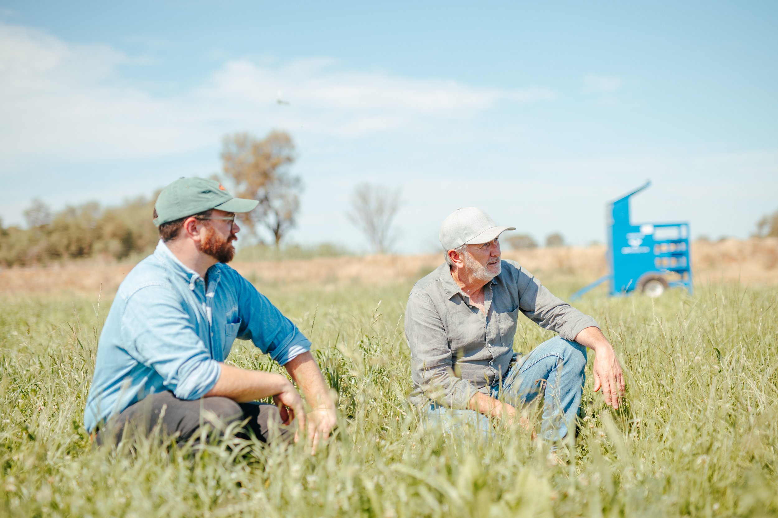 Two men sitting in a grassy field, engaged in conversation, with a blue piece of equipment or machinery in the background under a clear blue sky.