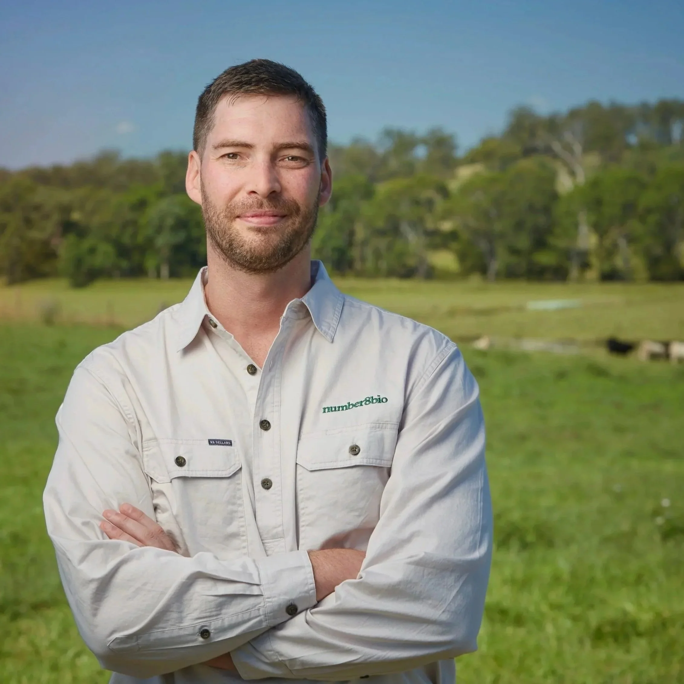 A man with short brown hair and a beard standing outside, wearing a white button-up shirt with a company logo that reads 'number8bio'. 