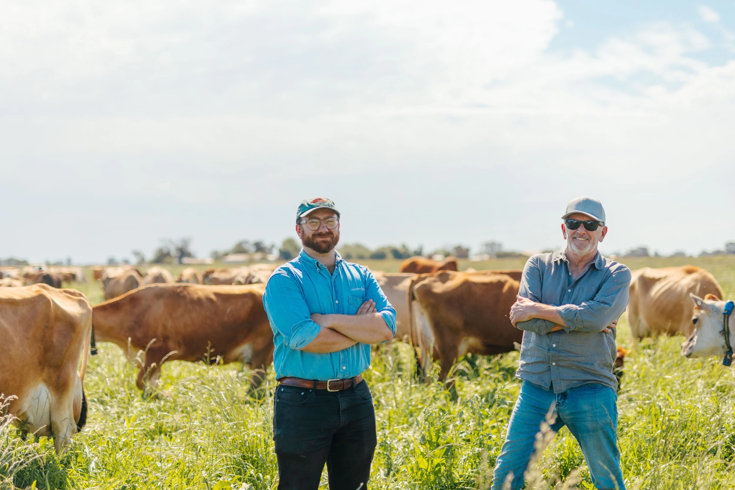 Two men standing in a field with cows, smiling and crossing their arms.