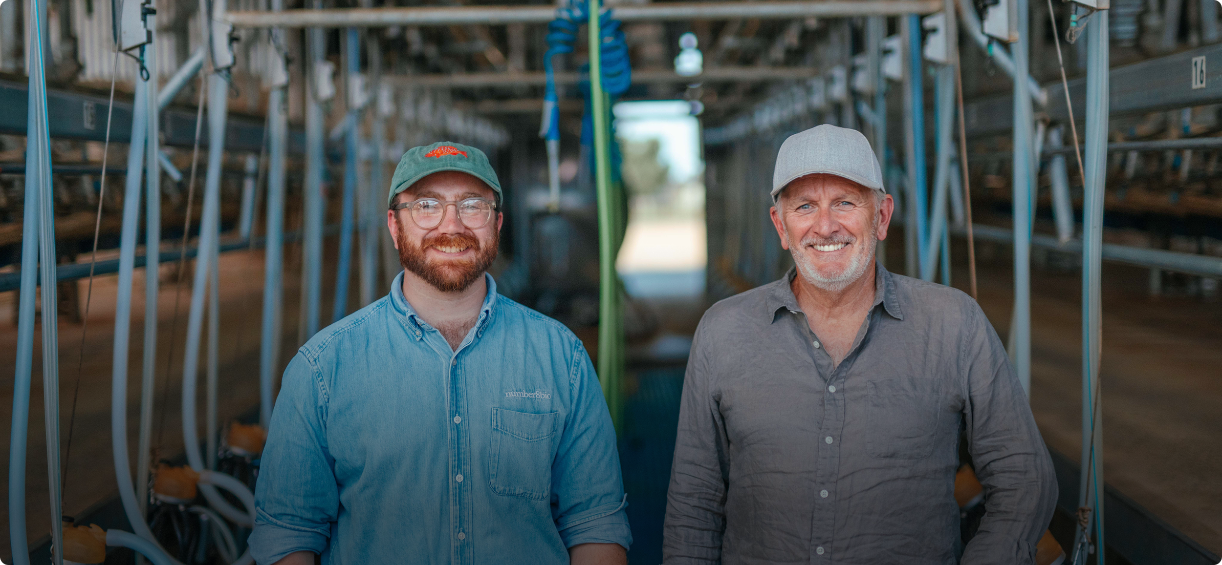 Two men standing inside a farm structure with metal shelving and equipment in the background, smiling at the camera.