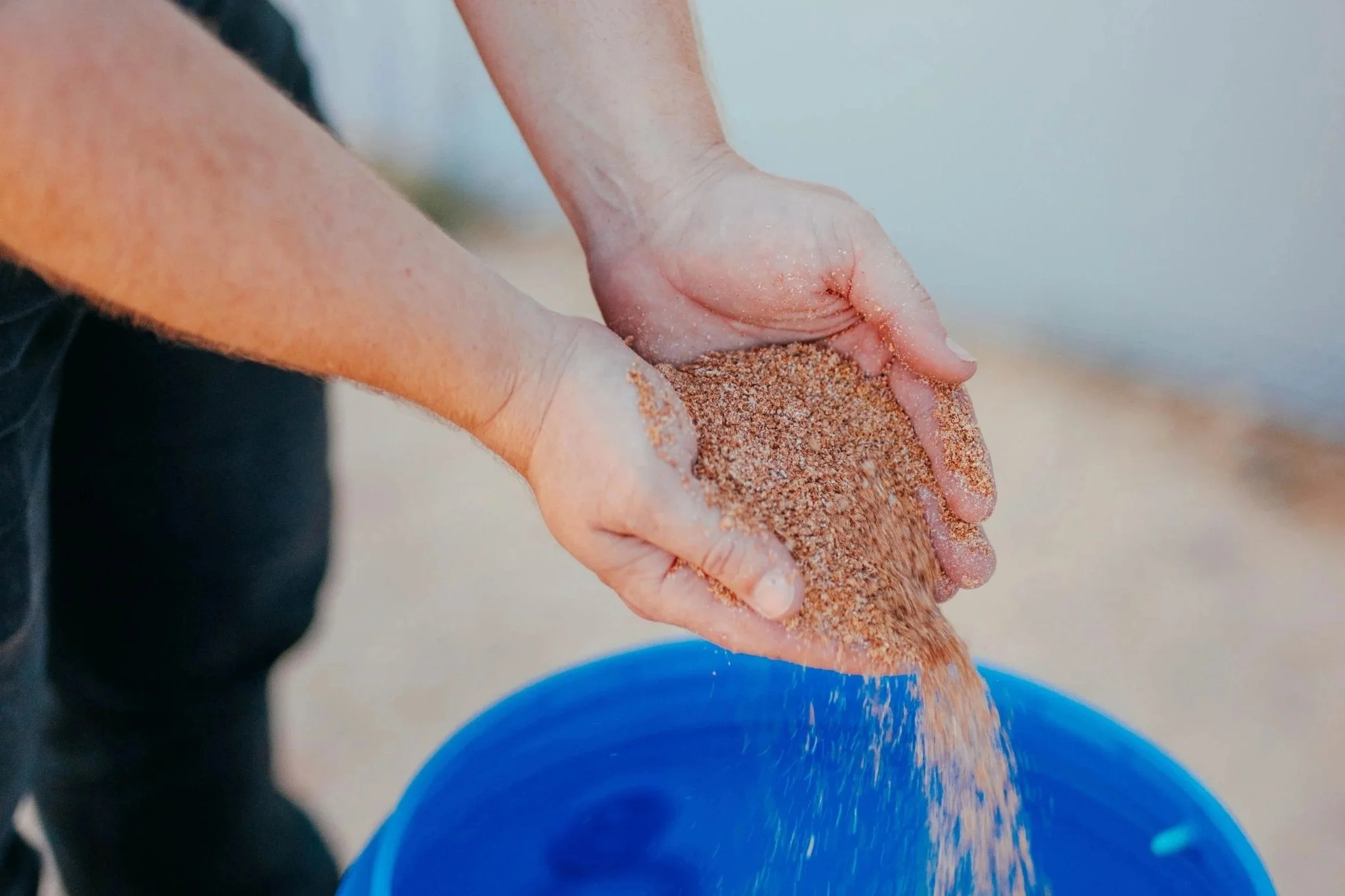Close-up of hands holding a handful of sand over a blue bucket.