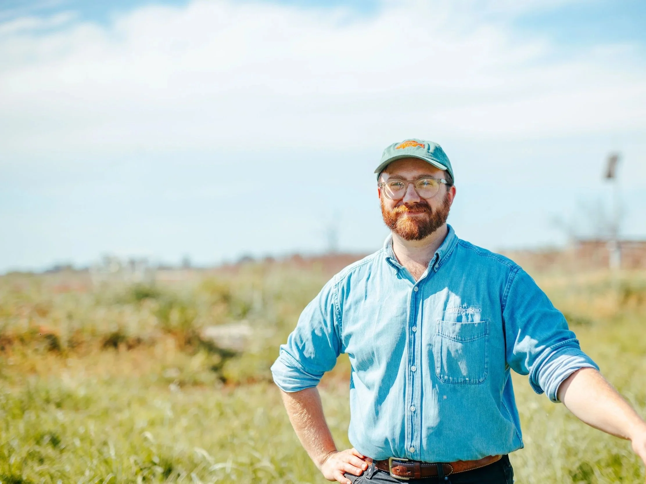 A man with a beard and glasses wearing a light blue denim shirt and a baseball cap, standing outdoors in a field with a cloudy sky in the background.