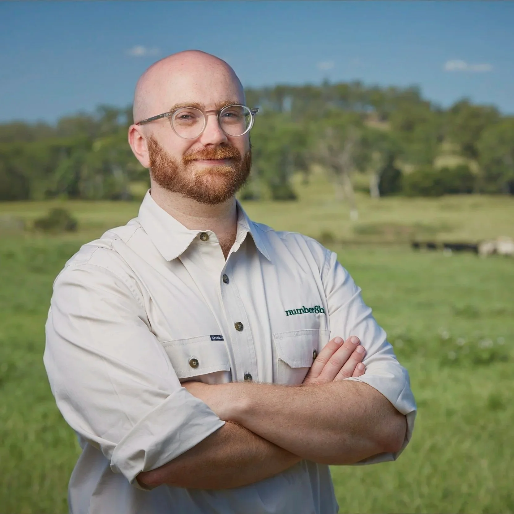 A man with glasses standing outdoors with his arms crossed, wearing a white button-up shirt, in a grassy field with trees and a blue sky in the background.