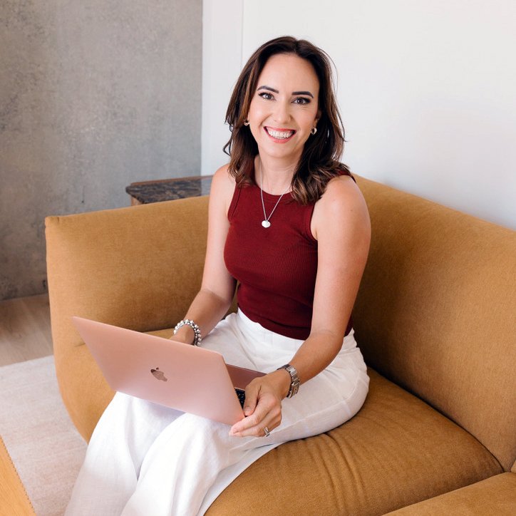 Cailie Ford smiling while working on a laptop, sitting on a mustard sofa