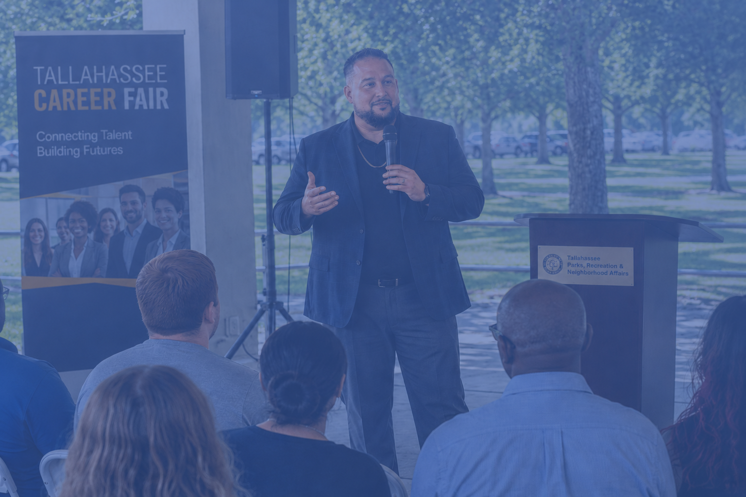 A man speaking at a Tallahassee Career Fair, standing at a podium with a microphone, in an outdoor setting with trees and parked cars in the background, while an audience listens.
