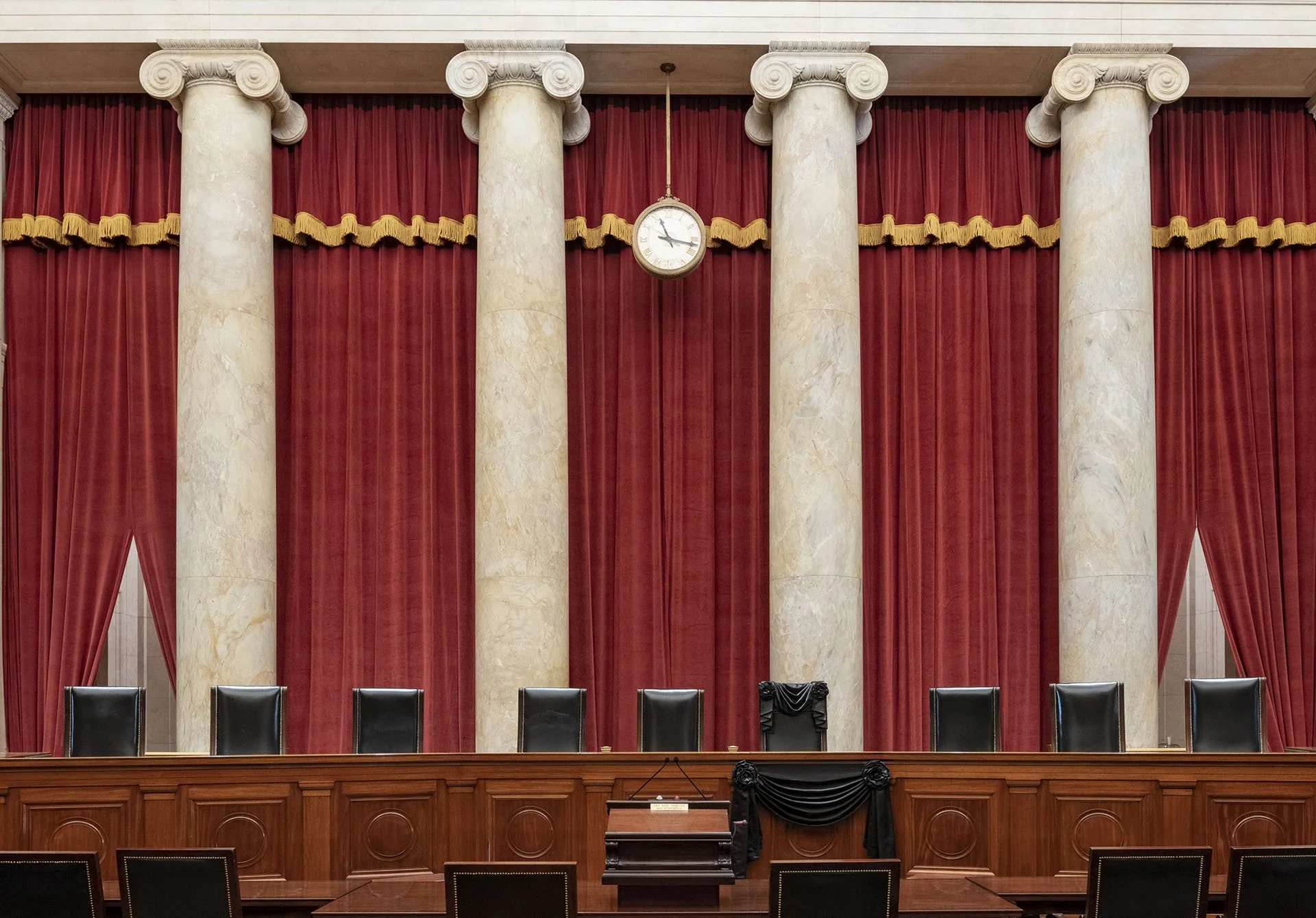 View of a formal government or courtroom setting with high red curtains, marble columns, a clock, and a wooden bench with chairs.