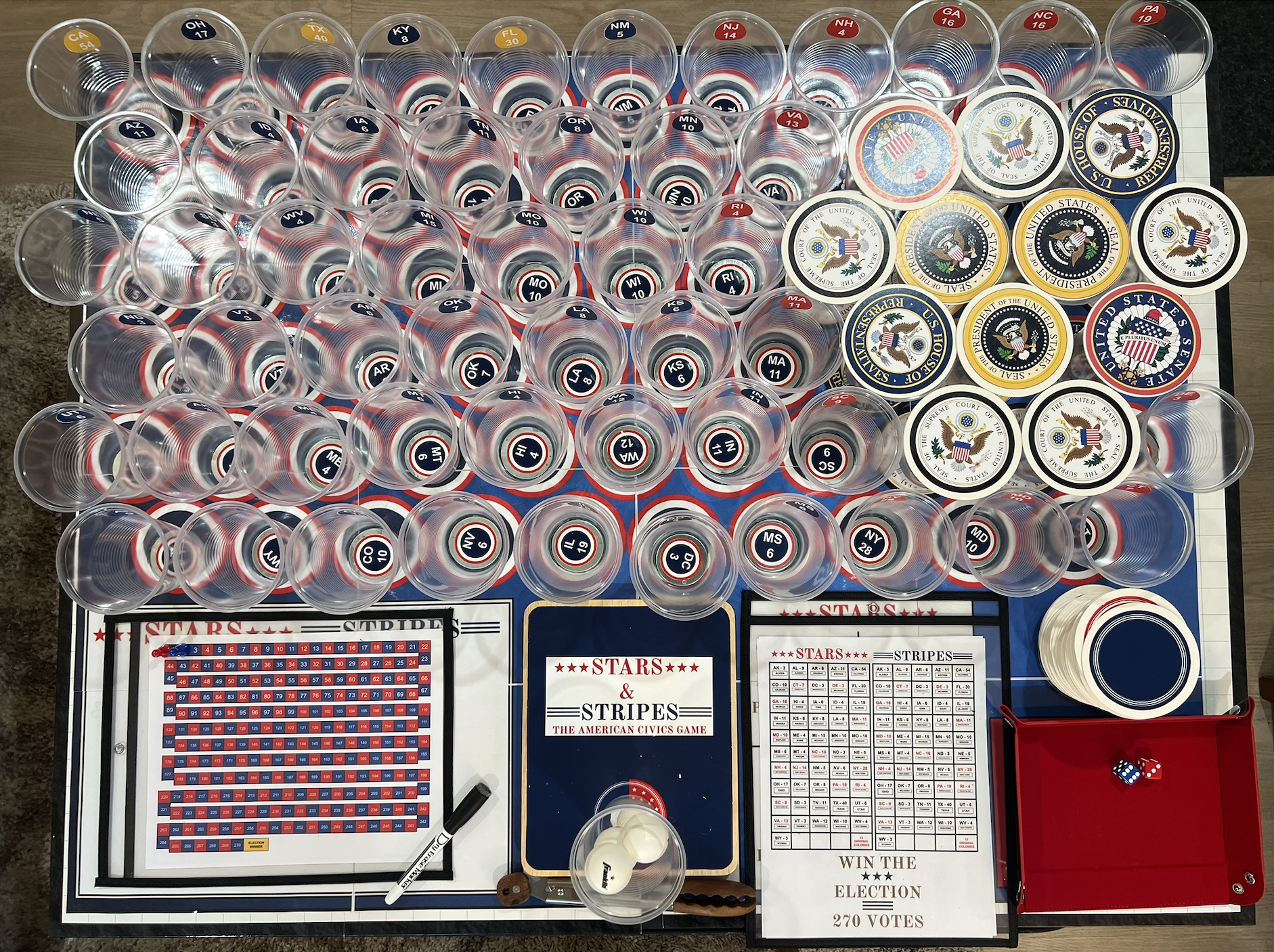A table setup for a political-themed game called 'Stars & Stripes,' featuring numerous clear plastic cups with state abbreviations and numbers, various badges with American symbols, a dry erase board for tracking points, and a red tray with dice.