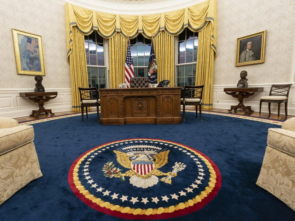 The Oval Office with a wooden desk, chairs, framed portraits, flags, and the presidential seal on the blue carpet.
