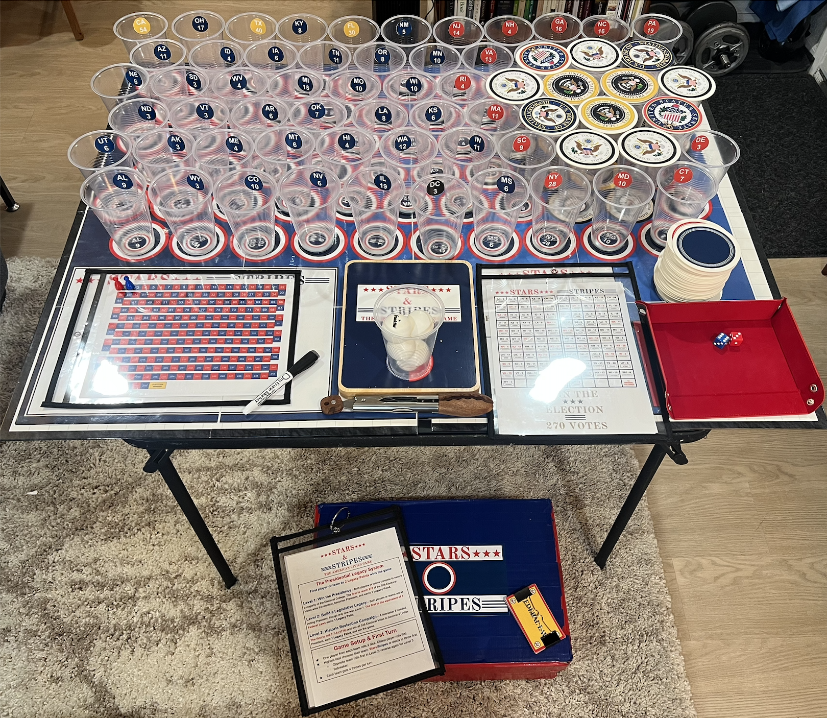 Table set up for a presidential election-themed game, featuring plastic cups with state abbreviations, stacks of chips, game boards, dice, and a rule sheet. The table has a red, white, and blue color scheme with patriotic labels and decorations.
