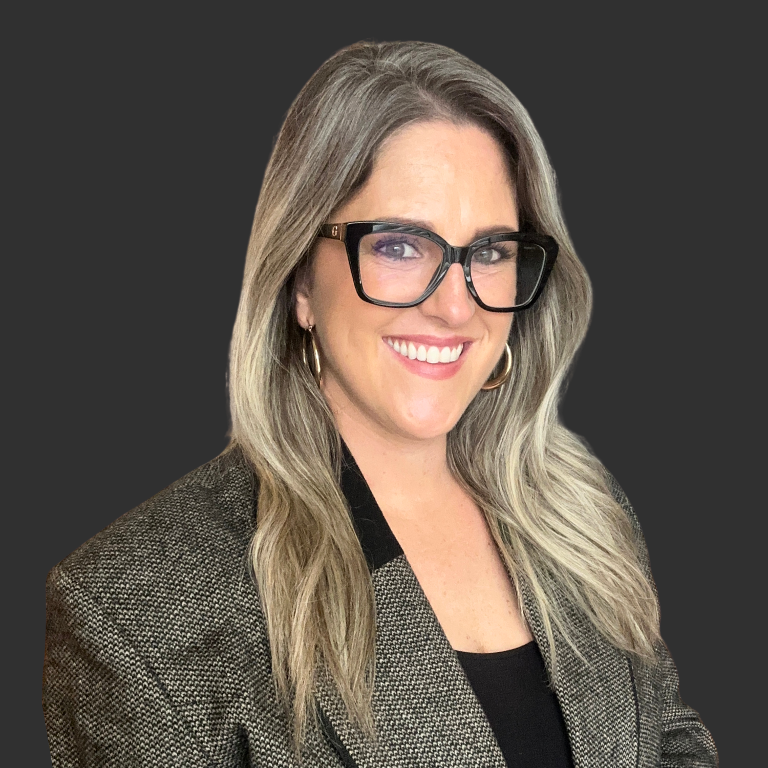 A woman with long, wavy blonde hair wearing black glasses, a brown checked blazer, black shirt, and gold hoop earrings, smiling at the camera against a dark background.
