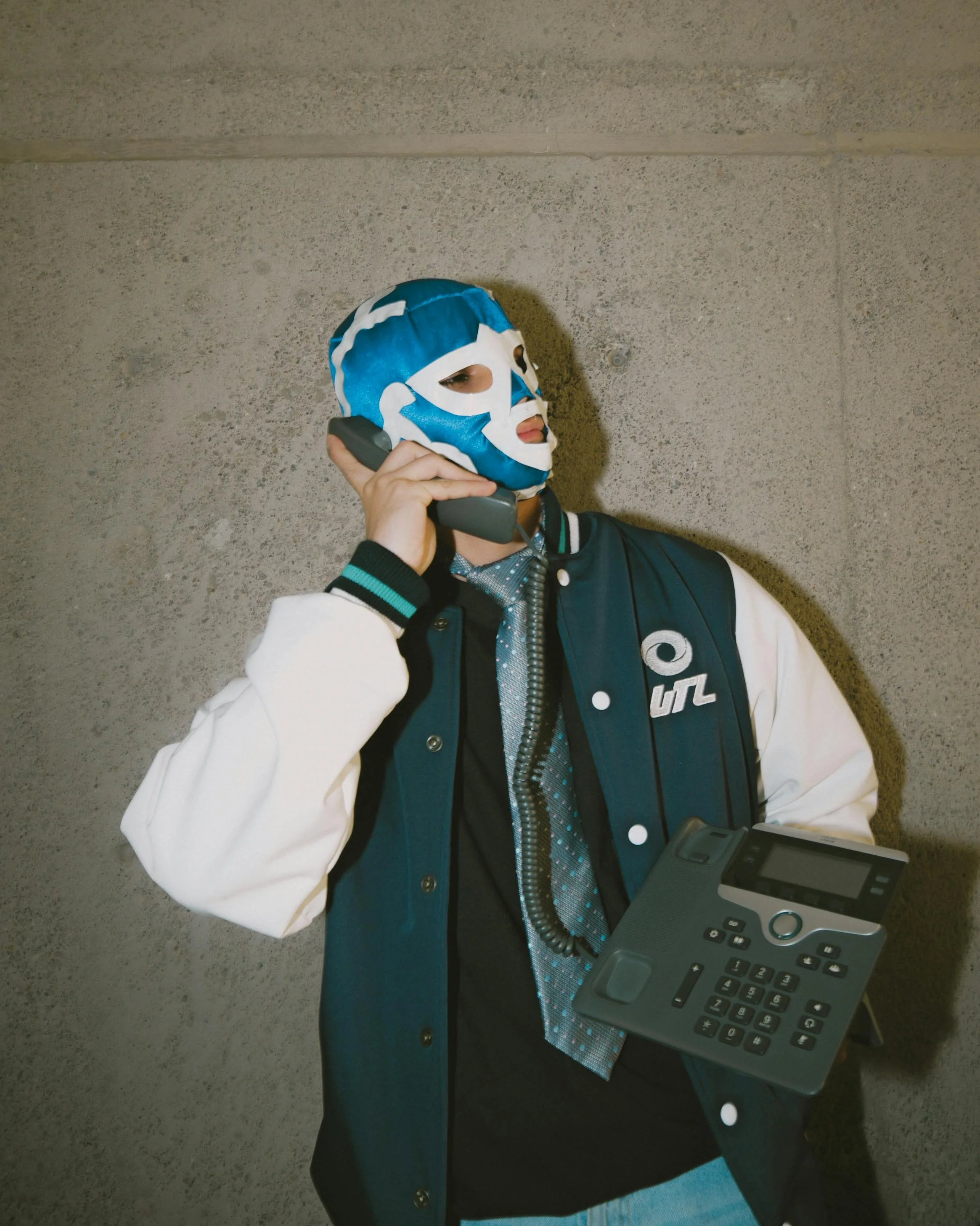 Person wearing a blue and white luchador mask, talking on a landline phone, holding a remote control, dressed in a varsity jacket, standing against a concrete wall.