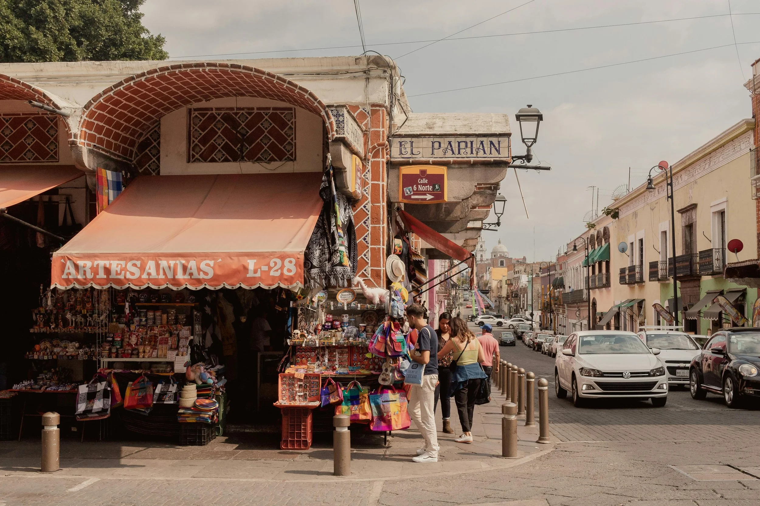Street scene with a small shop selling crafts and souvenirs, pedestrians walking along the sidewalk, colorful historic buildings, parked cars, and street lamps under a cloudy sky.