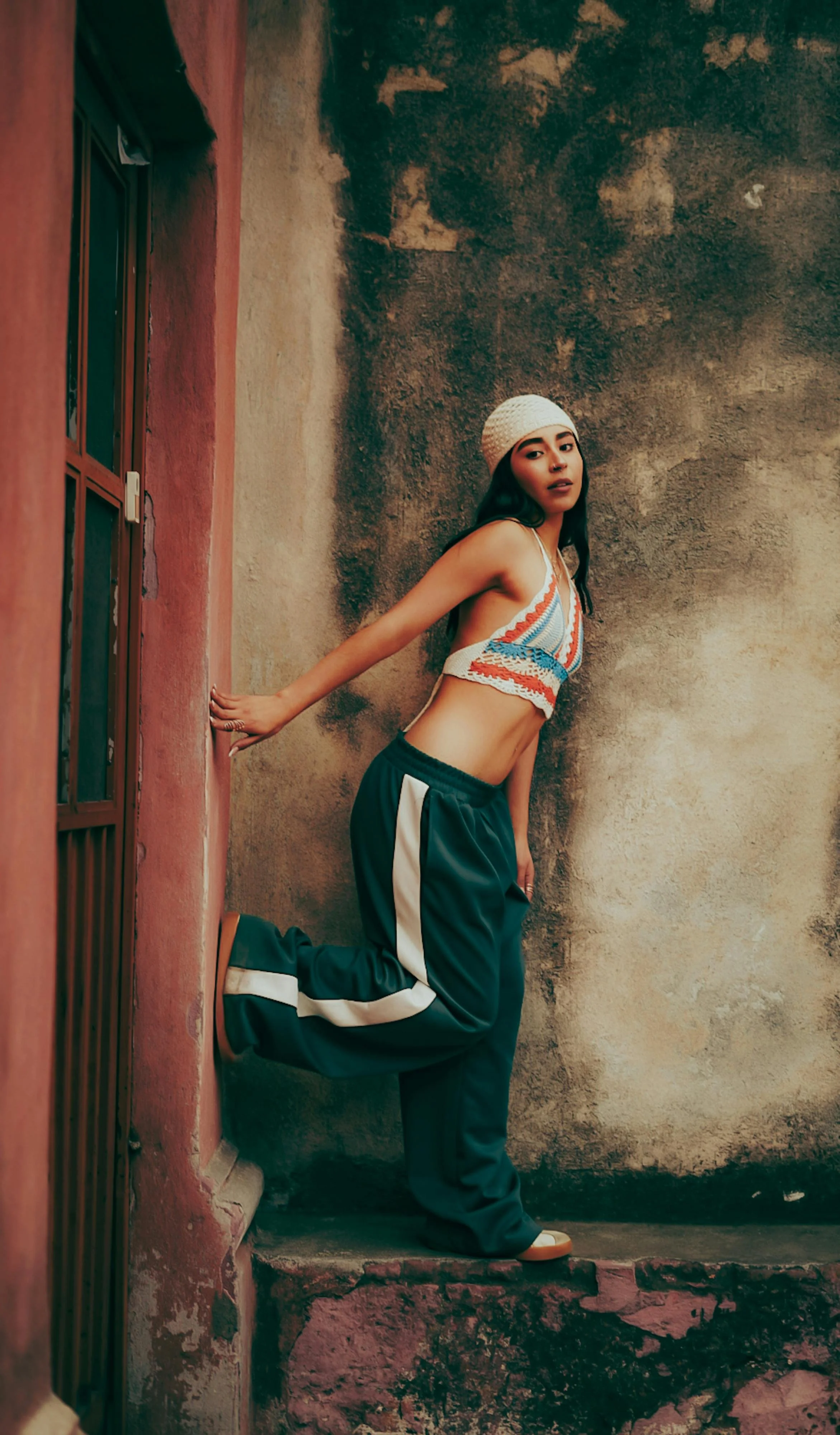 A young woman with dark hair wearing a white knit cap, a colorful crochet crop top, baggy dark green pants with white stripes, and sneakers, posing against an old, textured wall.