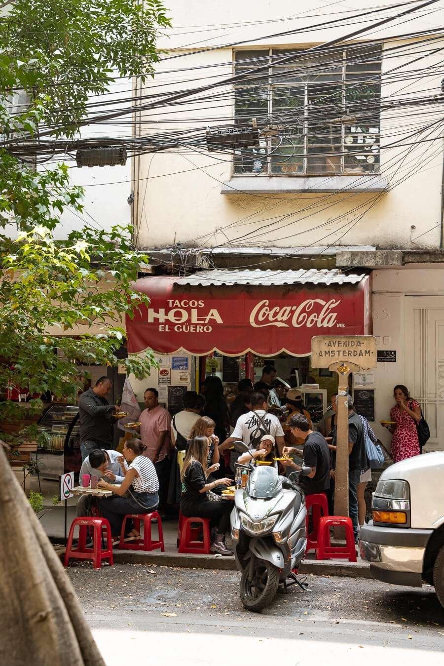 A busy small taco stand with a red Coca-Cola sign and a red awning that says 'Tacos Hola El Cuero' on a city street, with people eating and standing, a scooter parked in front, and an apartment building with exposed wiring above.