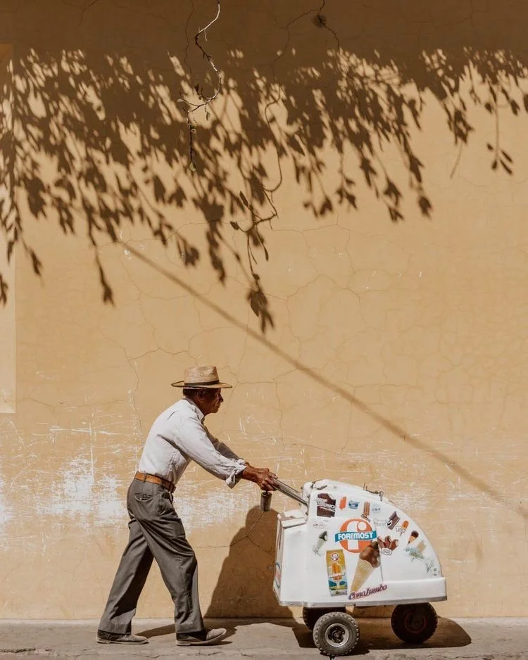An elderly man with a straw hat pushes an ice cream vending cart along a sidewalk. Shadows of tree leaves are cast on a yellow wall behind him.