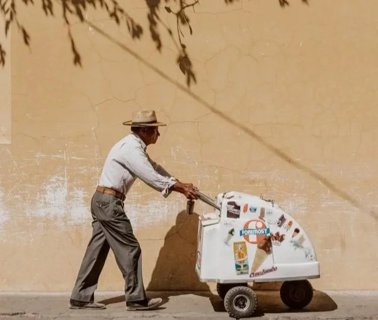 An elderly man in a hat is pushing a colorful ice cream cart on a sidewalk against a beige wall, with a shadow of tree branches overhead.