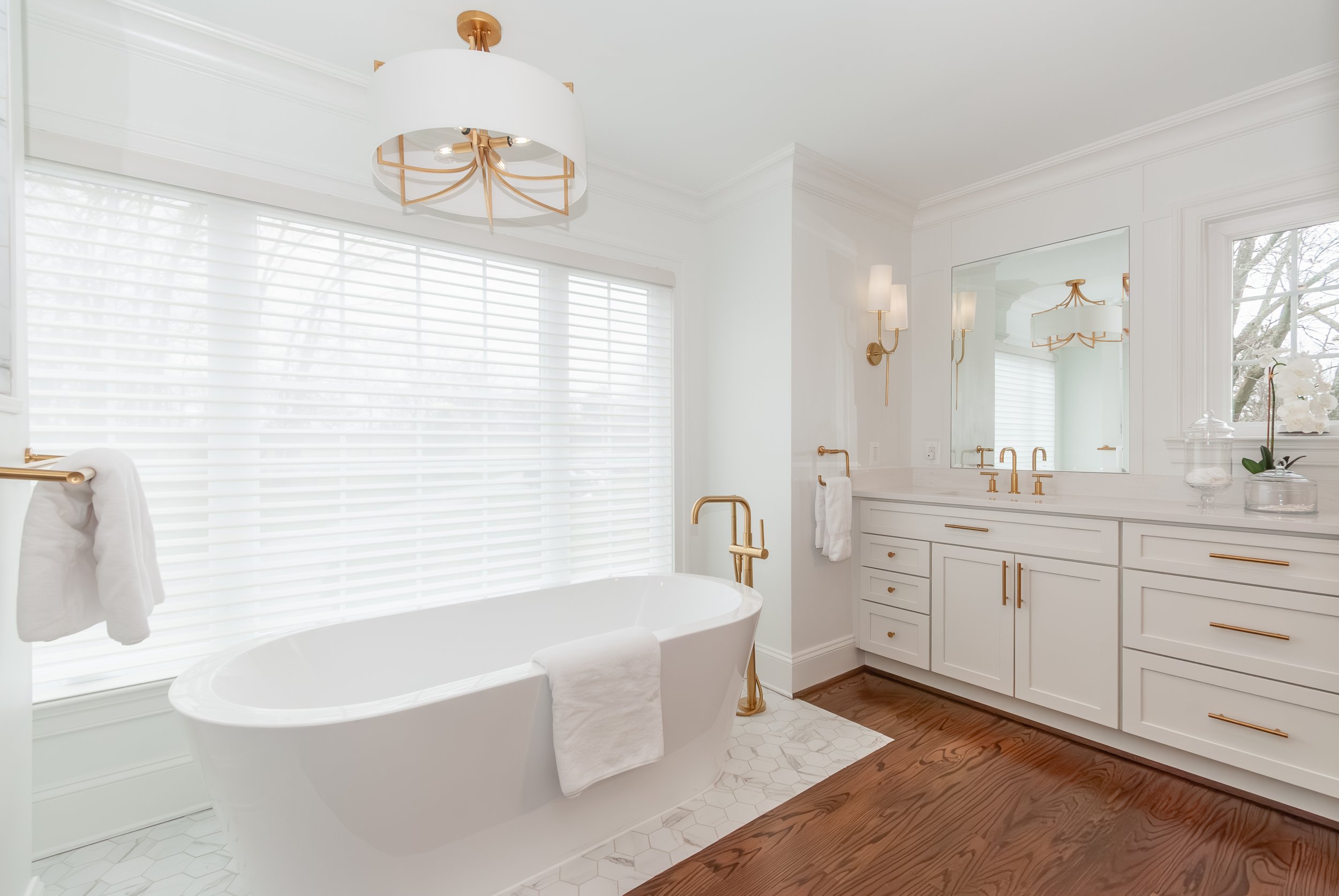Bright bathroom with white walls, bathtub, large window with blinds, wooden flooring, gold fixtures, and a white vanity with gold handles and a mirror.