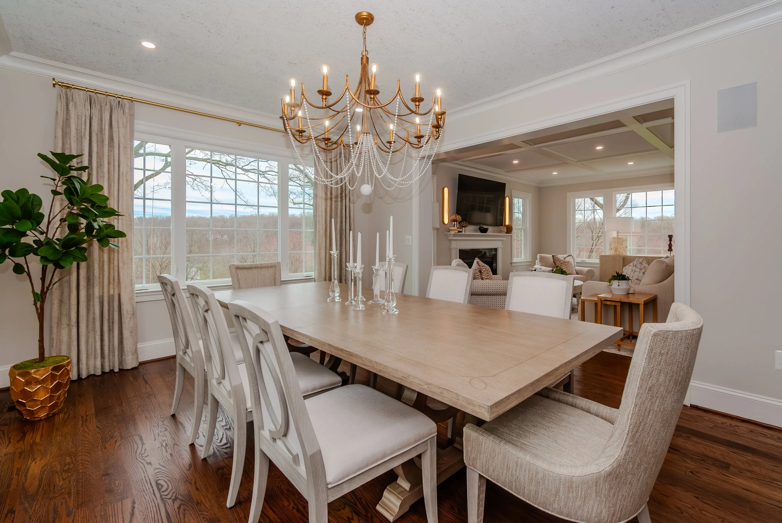 Dining room with wooden table and cream chairs, chandelier, large window with curtains, potted plant, and view of living room with fireplace and TV.