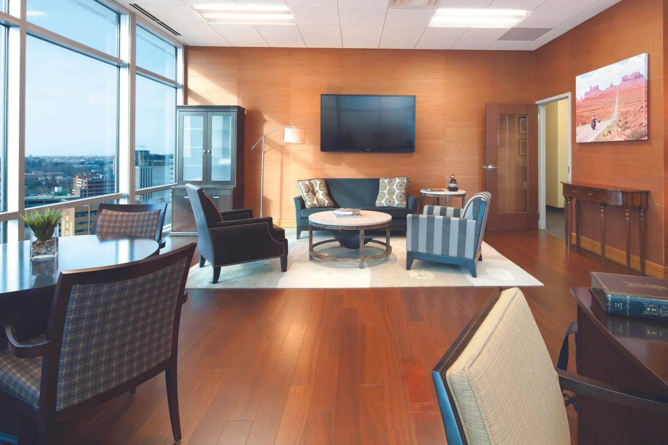 A modern hotel lounge with large windows, a flat-screen TV on the wall, a blue sofa, several armchairs, a coffee table, and decorative items. There is a city view outside the windows.