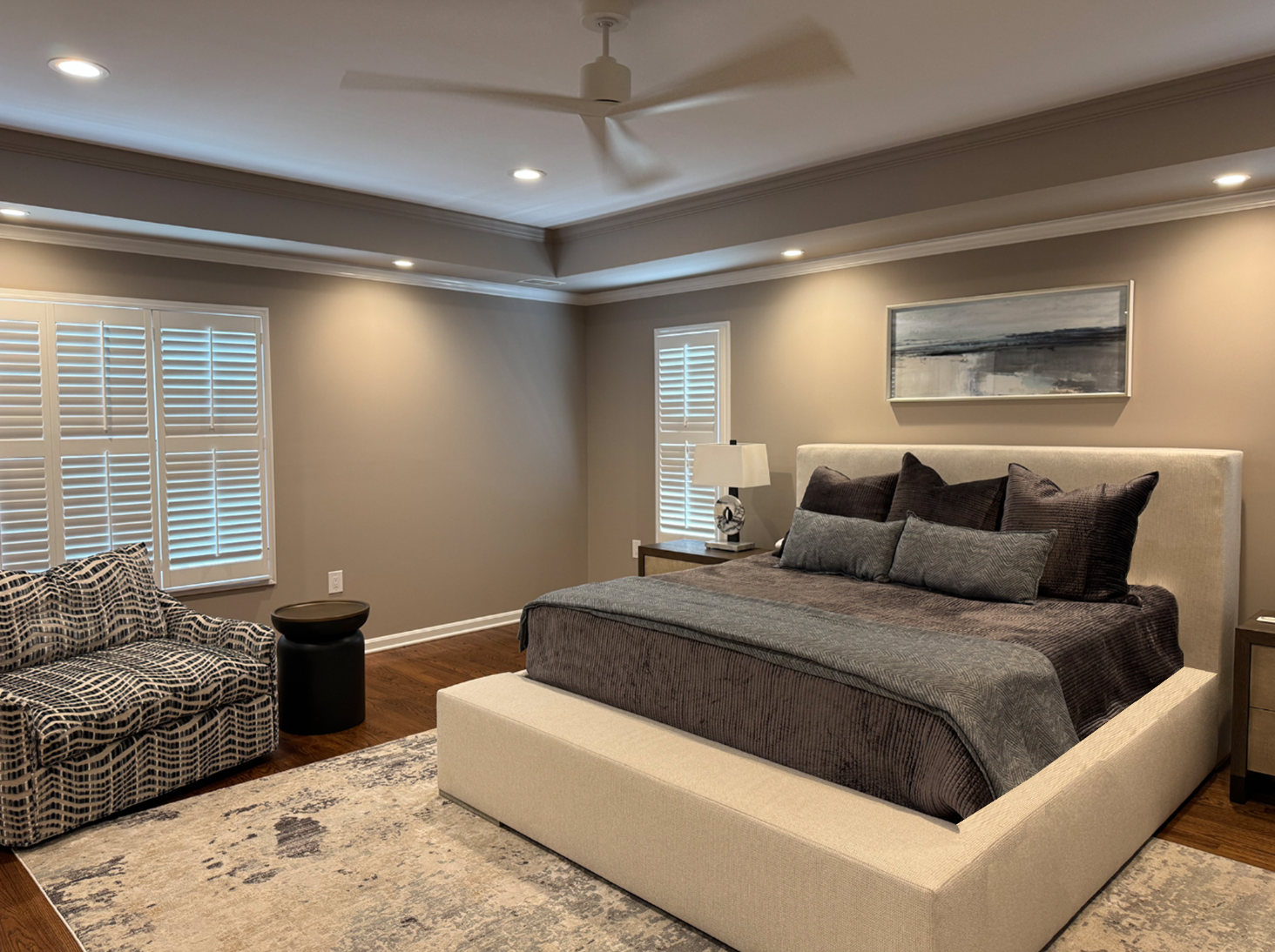Modern bedroom with a large bed, dark bedding, multiple pillows, a nightstand with a lamp, artwork above the bed, a patterned sofa, windows with white shutters, a ceiling fan, and recessed lighting.