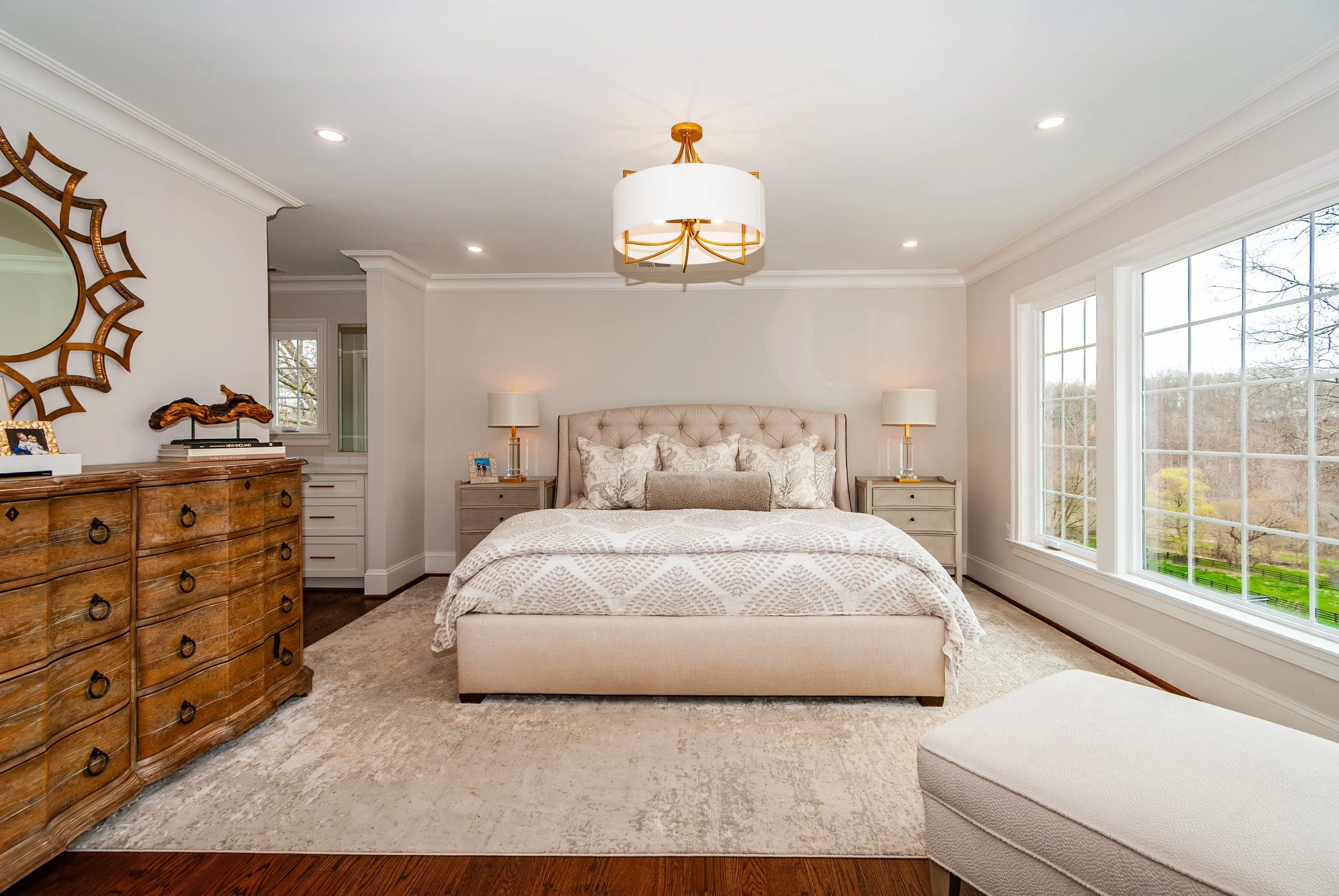 Bright, modern bedroom with large windows, beige upholstered bed, matching nightstands, and a wooden dresser, decorated with lamps, framed photos, and a decorative mirror.
