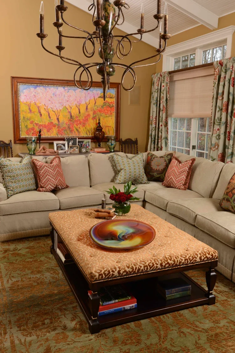 Living room with beige sectional sofa, decorative pillows, a wooden coffee table with a colorful glass bowl and a vase of red flowers, a large framed colorful painting of trees on a wall, a brass chandelier hanging from the ceiling, and windows with floral curtains.