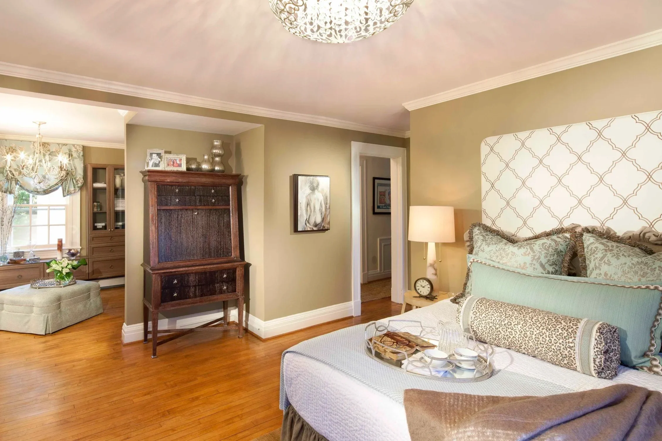 A cozy bedroom with soft lighting, featuring a bed with multiple pillows, a bedside table with a lamp and clock, and a hallway leading to other rooms. The room has hardwood floors, cream-colored walls, and decorative wall art.
